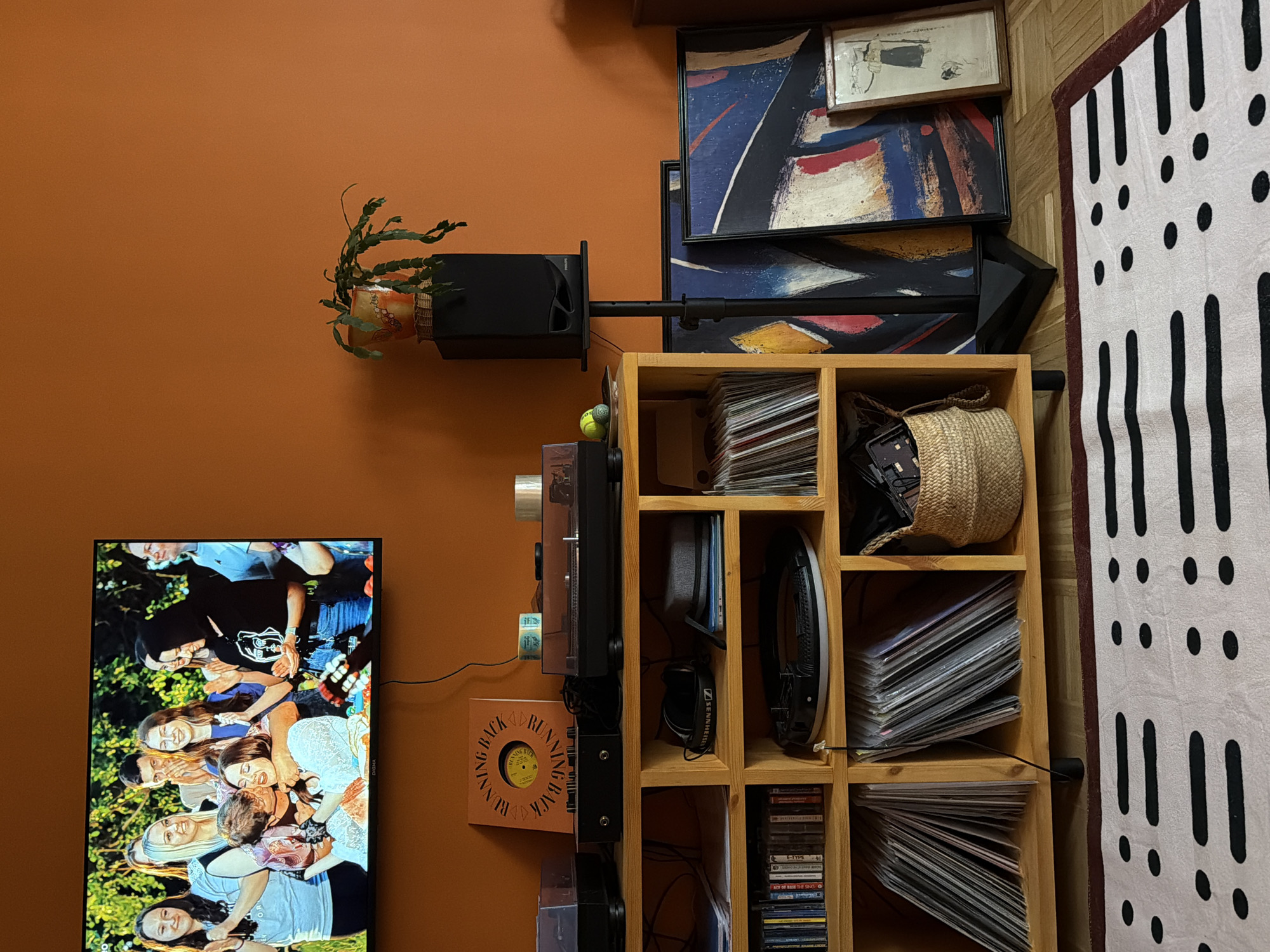 This image captures a warm and eclectic indoor living space, likely a living room, with a strong focus on music and art. The room features a prominent terracotta-colored wall and light wooden parquet flooring.
On the left, a large flat-screen television displays an outdoor scene of a joyful gathering of several people, including a child, with one person playing a guitar, suggesting a casual event or celebration. To the right of the TV, a sturdy light wooden shelving unit serves as an entertainment center, packed with various items. On its upper shelves, a leafy plant in a decorative pot sits next to a black Philips speaker mounted on a stand. A tennis ball, two record players (one black and one lighter-colored), and a woven basket filled with items are also visible. Many shelves are stacked with vinyl records and albums, some leaning. Headphones, with the "SENNHEISER" brand partly visible ("SENNHE" noted by OCR), are neatly placed on one shelf. The bottom shelves contain stacks of CDs and cassettes, with titles like "E-TYPE" and "FACE OF BASE THE SING" (likely "The Sign" by Ace of Base) discernible.
On the wall above the shelving unit, multiple framed artworks are stacked, including an abstract painting featuring bold strokes of blue, red, and yellow, and a smaller framed drawing or print. A white rug or tapestry with a black pattern of dots and vertical dashes partially covers the floor and leans against the unit on the far right. An album cover on the wall near the turntable has circular text that reads "RUNNING BACK." The overall aesthetic suggests a personal, lived-in space dedicated to leisure and cultural interests. The lighting is artificial, indicating an indoor setting. The location context provided is Khimki, Russia.