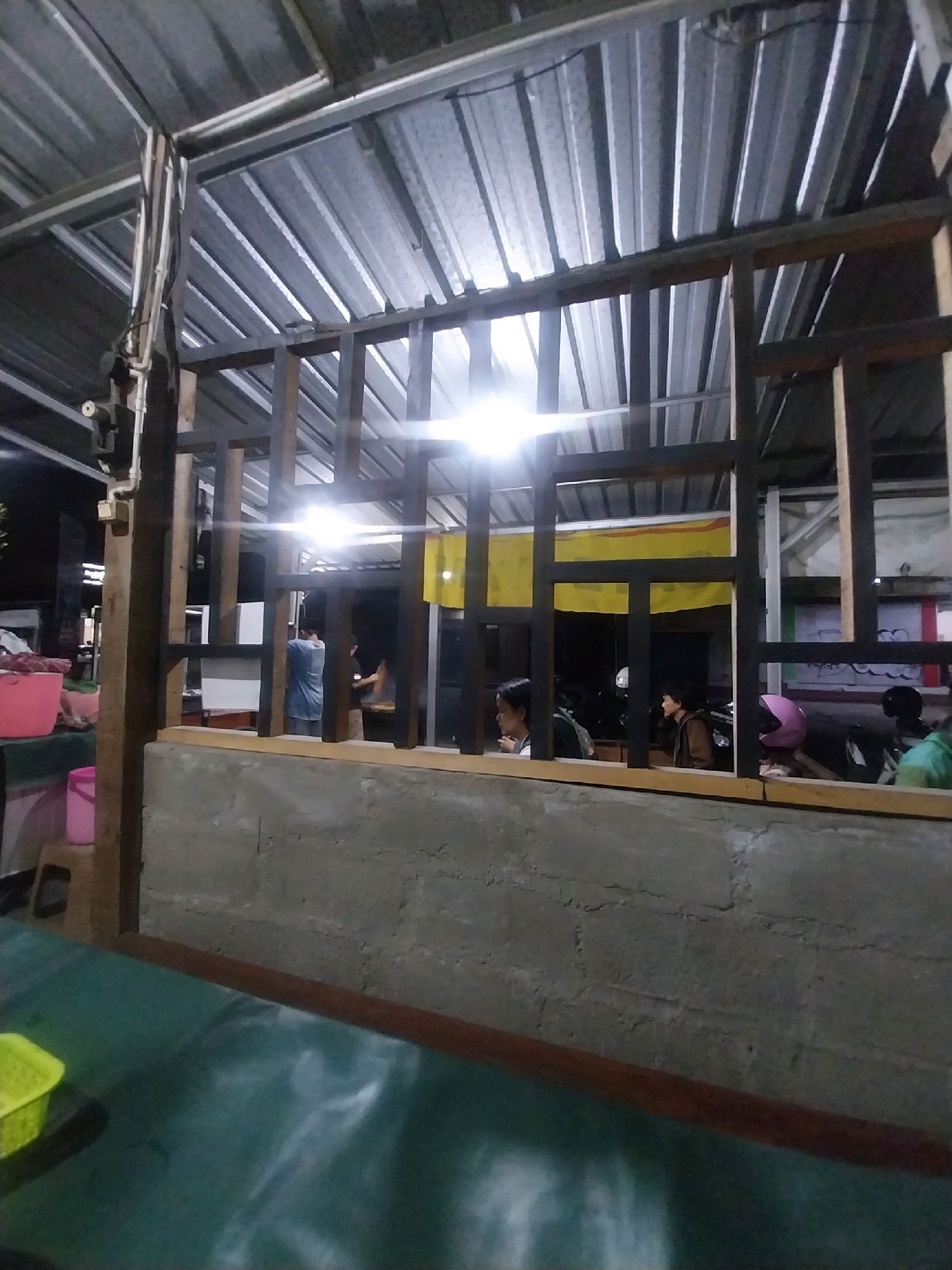 The image captures an interior view of an unfinished building, possibly a food stall or small establishment, in Trihanggo, Indonesia. The structure features a corrugated metal roof supported by metal beams and wooden framing. In the foreground, a rough concrete wall runs horizontally, with a wooden frame above it. Beyond the wall, several individuals are visible through the wooden lattice.
On the left, a person in a blue shirt stands near a white surface, possibly a counter or whiteboard. To their right, two individuals sit, facing towards the front of the establishment. One person wears a brown jacket, and another is partially visible. Further to the right, a person with a pink helmet is seated, and another person in a green shirt is visible in the background. Motorcycles are parked behind the seated individuals.
To the left of the wooden structure, pink buckets and a stool are visible, suggesting preparation or storage areas. The lighting appears to be artificial, with bright lights illuminating the interior and casting shadows. The overall scene depicts an active, albeit under-construction, commercial space.