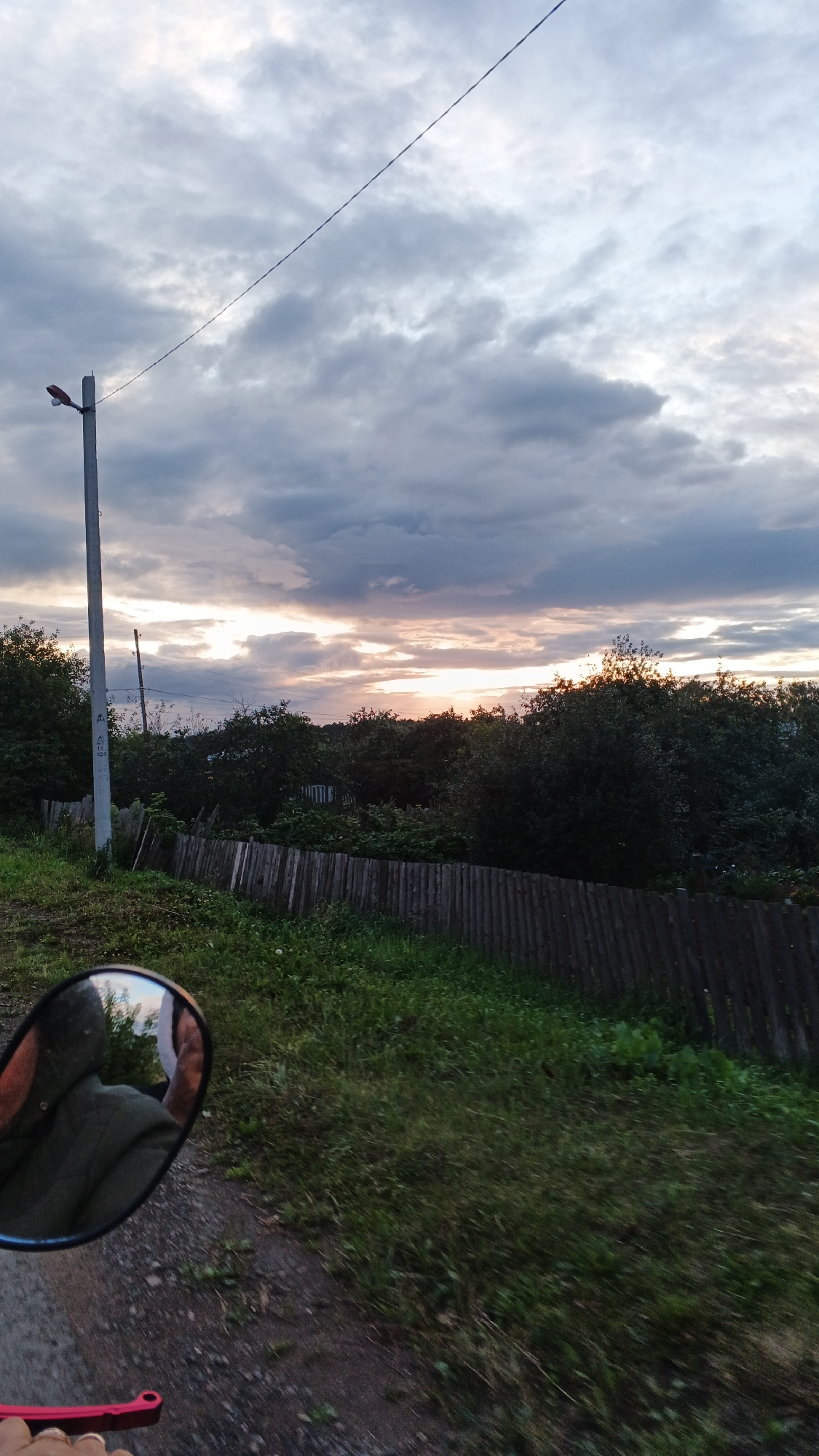 The image captures a cloudy sunset scene in a rural setting. In the foreground, a motorcycle's side mirror reflects a person wearing a green jacket and a white shirt. To the right of the mirror, a person's hand with a pink grip is visible. A dirt and gravel road runs along the bottom left of the frame, bordered by a wooden fence. Beyond the fence, lush green grass and trees line the landscape, with a pole and a wire stretching across the sky. The sky is filled with dramatic, grey and white clouds, with streaks of orange and yellow peeking through, indicating the sun is setting. In the background, dense foliage and trees obscure any further details of the environment. The location is identified as the village of Podgornoye, Russia, a small settlement likely characterized by such natural, uncultivated landscapes.