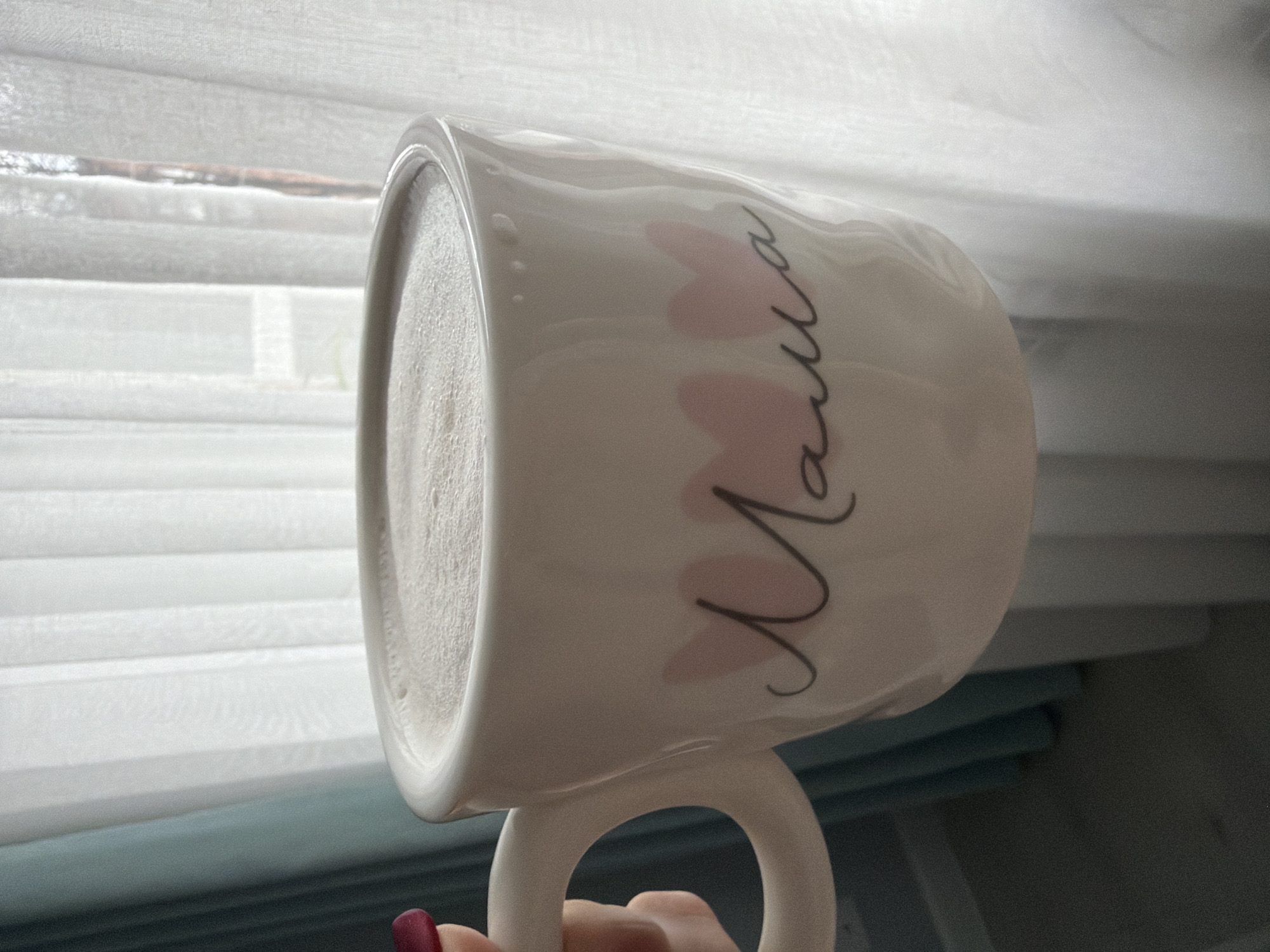 The image shows a white ceramic mug with the word "Mama" written on it in a decorative script with pink heart accents. The mug appears to be filled with a foamy, light-colored beverage, possibly coffee or tea with milk. A hand, identifiable by a fingernail painted in a dark shade, is holding the mug by its handle. The background consists of a window covered by semi-transparent blinds, obscuring the view outside. Based solely on the information in the prompt, it is impossible to determine the location is Votkinsk, Russia. There is no information in the image to connect it to that city.