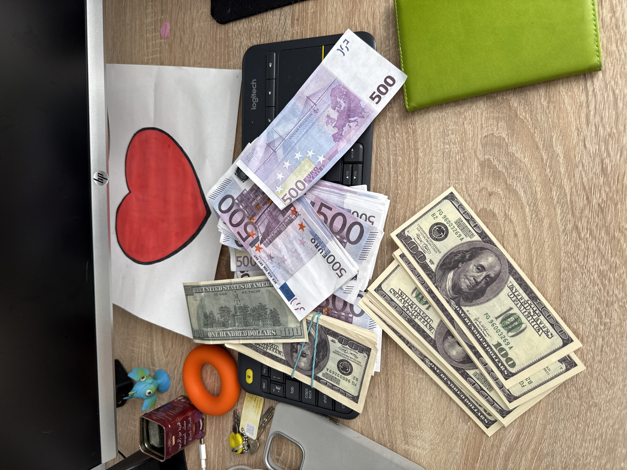 A desk contains a Logitech keyboard with stacks of 500 Euro notes and bundled stacks of US 100 dollar bills. Additional items on the desk include a drawing of a red heart, a green wallet, a monitor, a blue plastic bird, an orange ring, a small tin, a grey cell phone case, and keys. The setting is not identifiable as Rostov-on-Don, Russia.