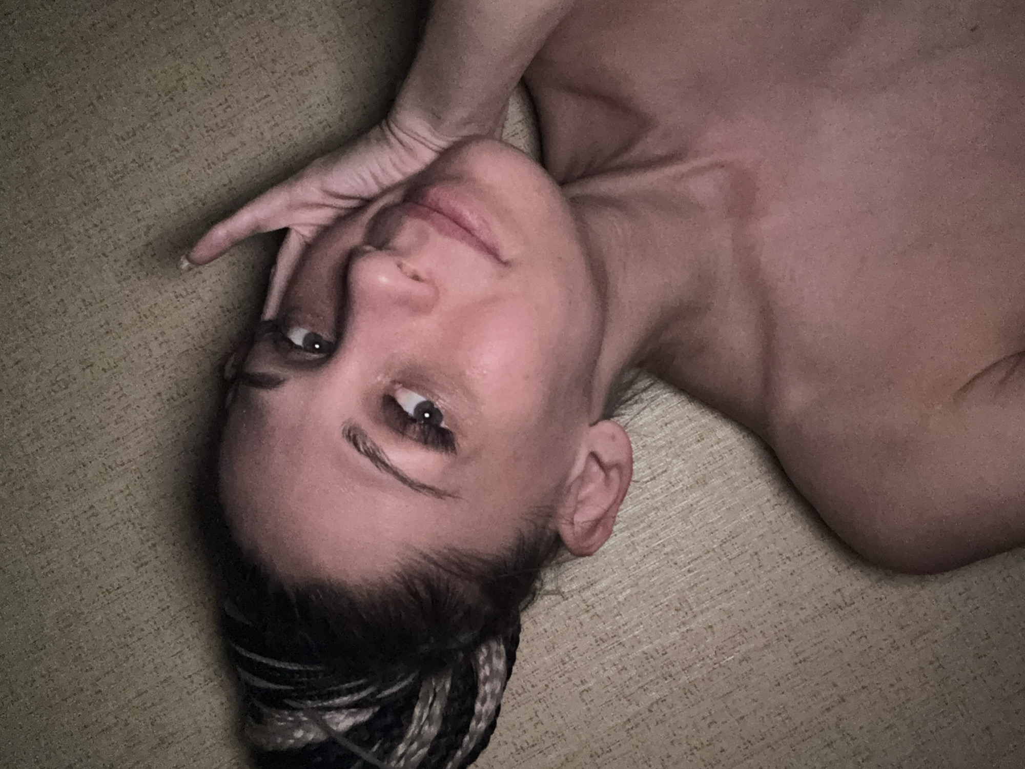 A woman with braided hair is lying on a beige textured surface. Her head is tilted to the side, and her hand is resting near her face. She is looking directly at the camera. Her skin tone is fair, and her eyes are open. Her upper body is exposed.
There is no information to support this image being in the city of Rusanovo, Russia.