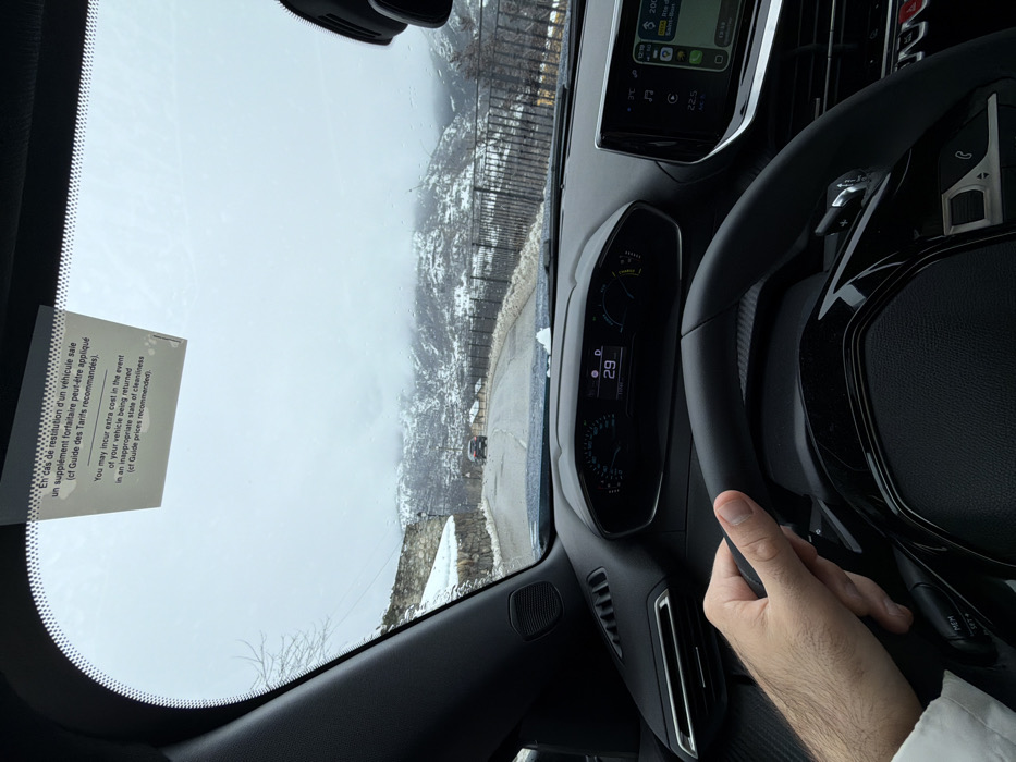 The scene depicts a first-person view from the driver's seat of a vehicle traveling on a winding, partially snow-covered mountain road in Courchevel, France. The navigation system visible on the infotainment screen indicates "Rte de Saint-Bon," confirming the location within the ski resort area.
**Vehicle Interior:**
The immediate foreground shows the dashboard and steering wheel of a modern car, likely a Peugeot. A right hand, wearing a white cuff on the forearm, grips the black steering wheel at approximately the 3 o'clock position. The digital instrument cluster displays "29" (likely speed in km/h or possibly temperature), and the infotainment screen on the right shows a navigation map with "200m Rte de Saint-Bon," a time of "10:18," and an interior temperature of "22.5°C." A rectangular paper notice is affixed to the upper center of the windshield, containing text in both French and English regarding potential cleaning surcharges for rental vehicle returns.
**Exterior Environment:**
Beyond the windshield, an overcast, cloudy sky dominates the upper portion of the frame. The road ahead is asphalt, appearing damp or slightly wet, with patches of snow and slush accumulating on its edges. The surrounding landscape consists of snow-covered mountains and hillsides. Sparse, bare deciduous trees are visible on the left. A low stone retaining wall lines the left side of the road in the mid-ground, while a dark metal fence or barrier runs along the right side, overlooking a downward slope. Scattered buildings, characteristic of chalets or mountain dwellings, are visible among the snow on the right-hand hillside in the distance. Another dark-colored vehicle (possibly an SUV) is observed ahead on the road, traveling in the same direction.