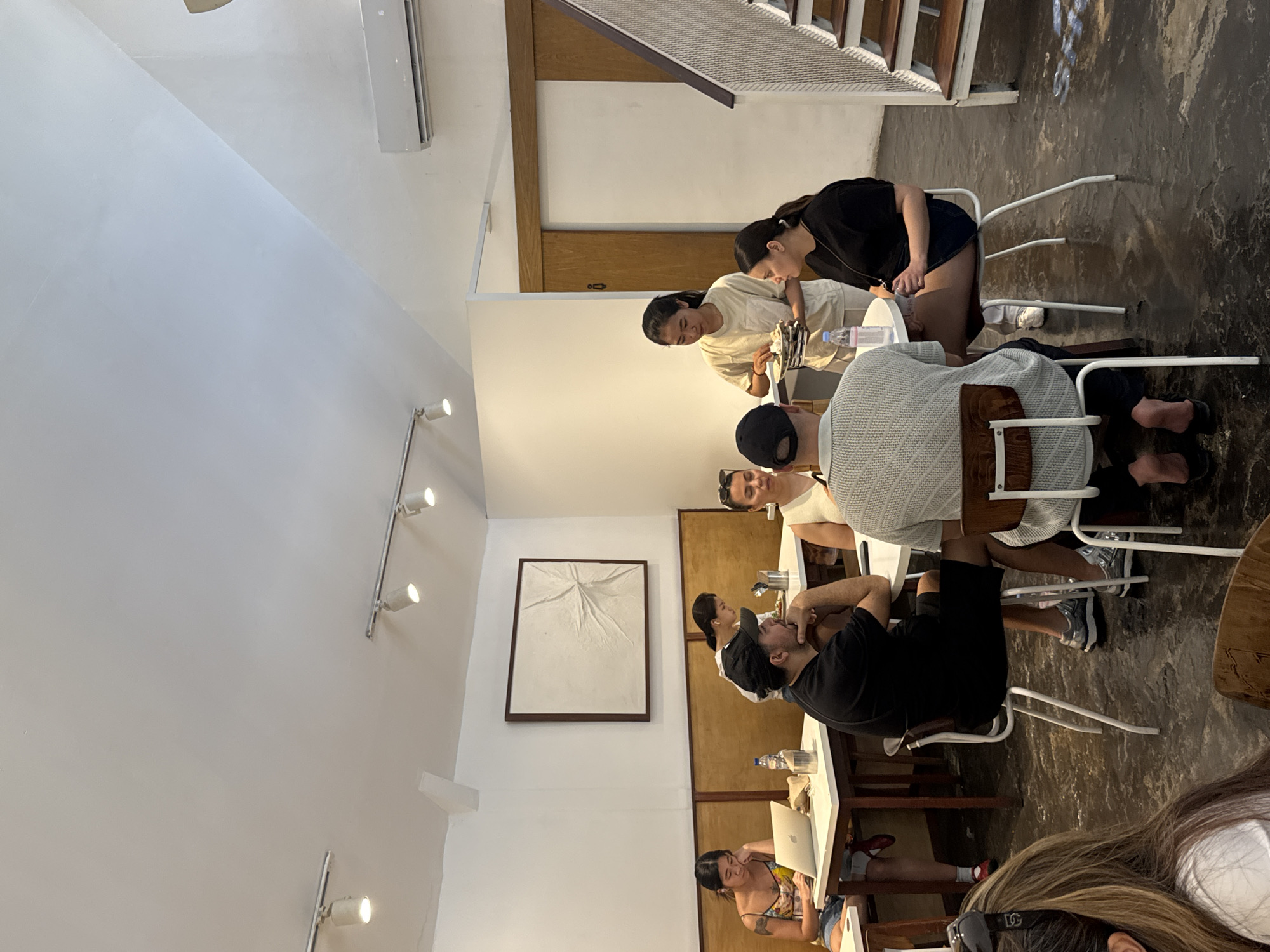 The image shows the interior of a restaurant in North Kuta, Indonesia. A group of people are sitting at a long table. Some are engaged in conversation, while others are working on laptops. The restaurant has a modern, minimalist design with white walls and wooden accents. There is artwork hanging on the wall. The floor is concrete.