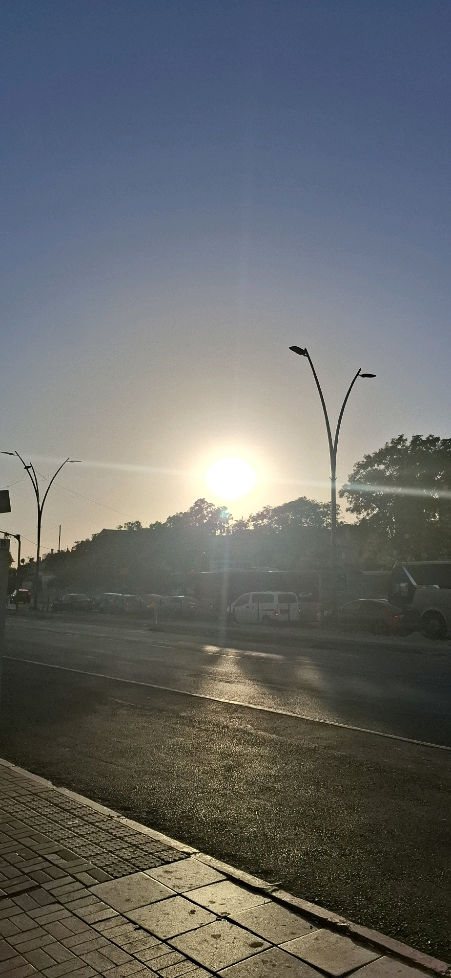 The image captures a street scene in Be'er Sheva, Israel, during sunset. The sun is low in the sky, casting a warm, golden light and creating lens flare. In the foreground, a paved sidewalk runs alongside a wet asphalt road, indicating recent rain or dew. Traffic lights and street lamps are visible, with two distinct lamp posts silhouetted against the sky. Several vehicles, including vans and cars, are present on the road, suggesting moderate traffic. Trees and buildings form the backdrop, partially obscured by the bright sunlight. The overall atmosphere is serene, with the setting sun creating a dramatic and visually striking effect on the urban landscape.