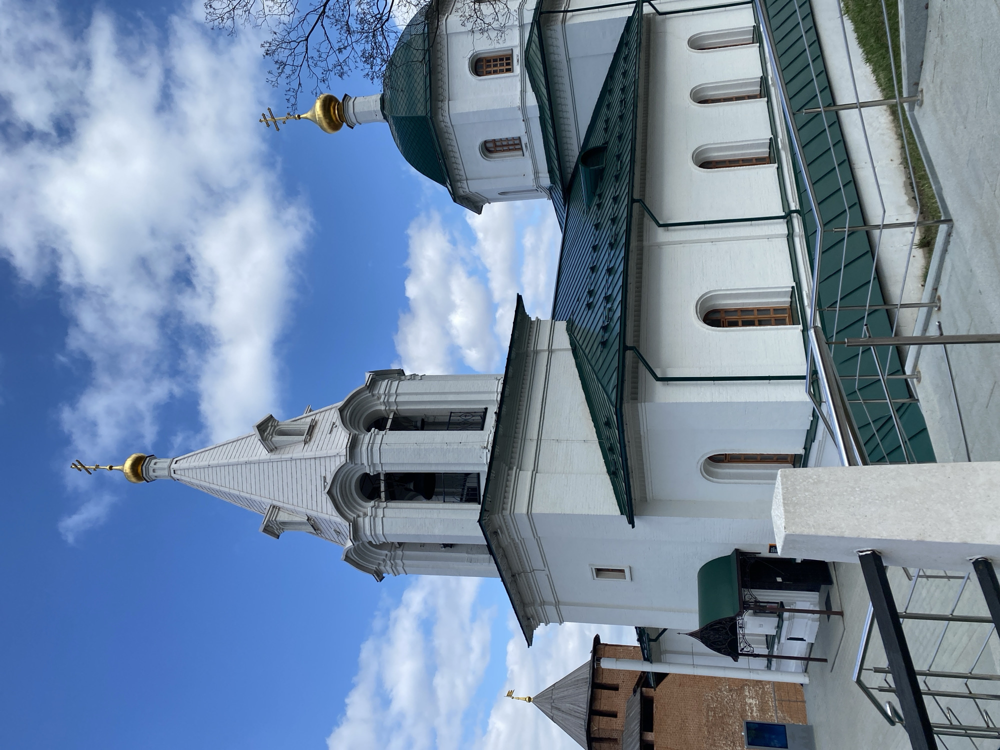 This image captures a vibrant architectural complex, likely a Russian Orthodox church or monastery, set against a bright blue sky dotted with scattered white clouds. The primary subject is a large, pristine white building with distinctive dark green roofs.
On the left side of the frame, a tall, elegant white bell tower (or spire) rises, featuring multiple tiers with arched openings, suggesting a belfry, and culminating in a golden onion dome topped with a traditional Orthodox cross. To the right of the bell tower, another section of the church features a larger golden onion dome, also crowned with a cross, typical of Russian ecclesiastical architecture. The white walls of the buildings are punctuated by several arched windows with wooden frames.
In the foreground on the right, a modern metal railing suggests a pathway or viewing platform, with a small patch of green grass visible beyond it. Below the main church structure and to the lower right, a portion of an older, reddish-brown brick building is visible, likely a historical wall or tower, which aligns with the context of a city like Nizhny Novgorod, Russia, known for its kremlin. Bare tree branches are seen in the upper left corner against the sky, indicating a season like early spring or late autumn. The clear skies and bright illumination suggest it is daytime. No people or specific events are depicted, and no visible text is discernible.