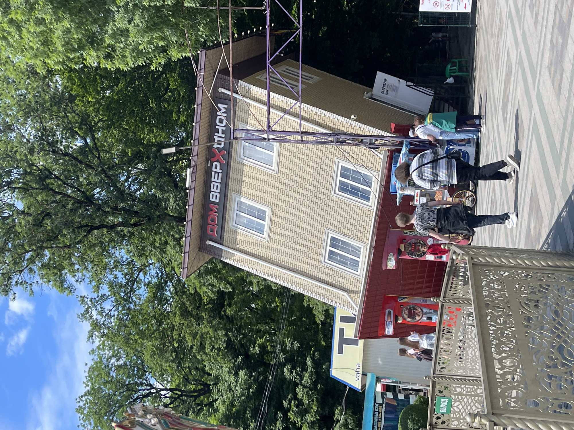 The image shows an outdoor scene in a park or amusement area, featuring a prominently displayed upside-down house attraction. The house is a two-story structure with yellow brick siding and white-framed windows. The words "ДОМ ВВЕРХ ДНОМ" are visible on the upper part of the house, meaning "upside-down house." A metal support structure is attached to the house.
In the foreground, a paved walkway with a geometric pattern is visible. A decorative metal railing with ornate designs runs along the left side of the walkway. Several people are walking on the paved area. Two individuals, a person wearing a black and white striped shirt and another in a dark t-shirt, are walking away from the camera. A young person in a dark t-shirt is walking towards the camera. Further back, a group of three people, two women and a man, are standing near a food stall.
Several smaller structures and attractions are visible behind the upside-down house. To the left of the house, there's a sign with the letter "T" and the word "vaha." Adjacent to this is a red structure that appears to be a game or attraction, labeled "BOXER." Another similar attraction is visible to the right. A food cart with "Pepsi" branding is also present.
The environment is characterized by lush green trees surrounding the area, suggesting a sunny day with a blue sky visible in the background. The overall impression is of a leisure or entertainment venue.
The location is identified as being in the city of Stavropol, Russia.