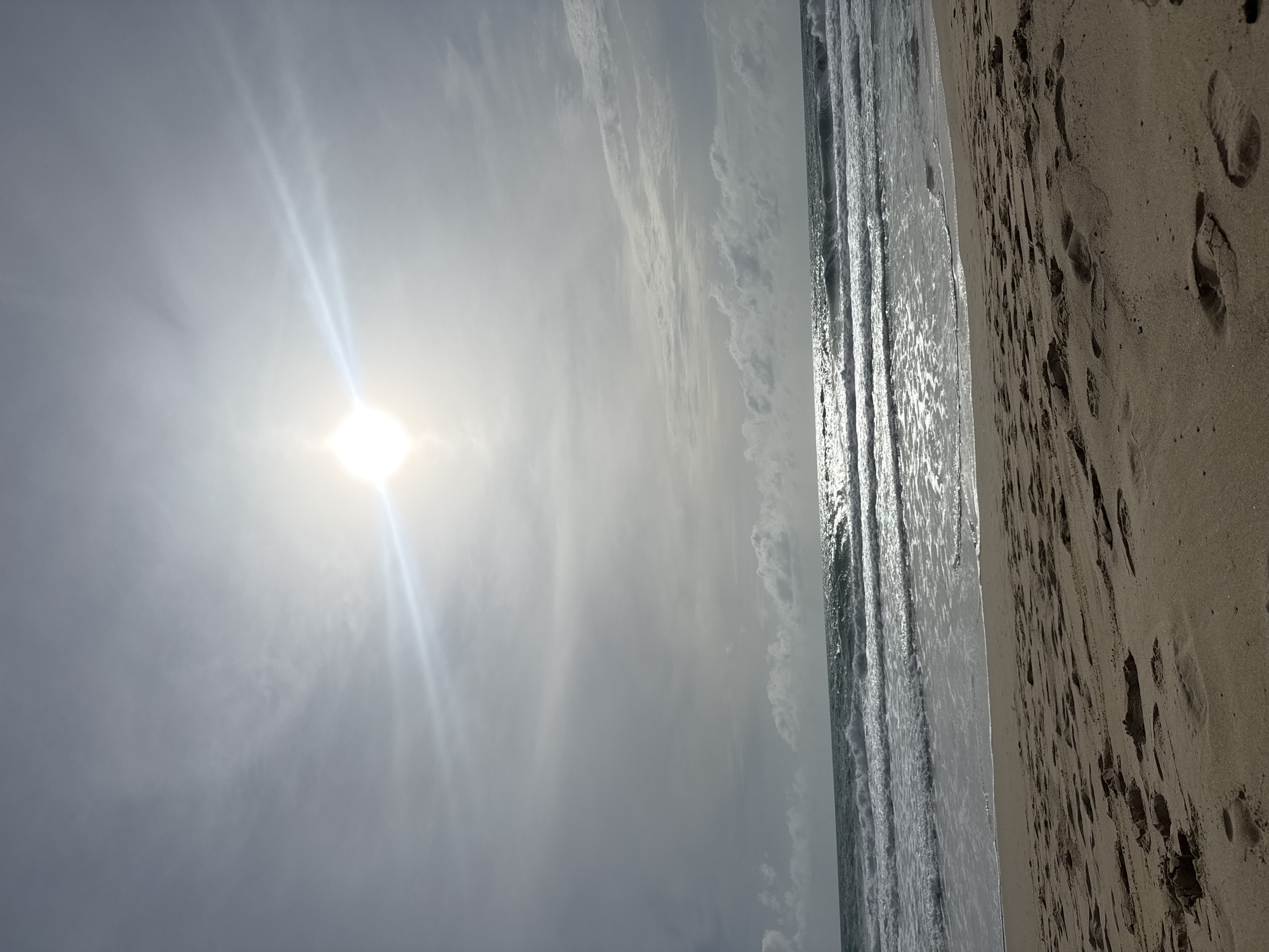 The image depicts a beach scene in Choeng Thale, Thailand, under a bright, overcast sky. The sun is visible as a strong, diffused light source in the upper portion of the frame, casting a glare and lens flare across the sky and the ocean surface. The sea is characterized by moderate waves with white crests, creating a shimmering effect as they reflect the sunlight. The shoreline is sandy, with visible footprints imprinted in the wet sand closest to the water and drier sand further up. No people are present in the frame. The environment is a typical coastal setting with sand, sea, and sky.