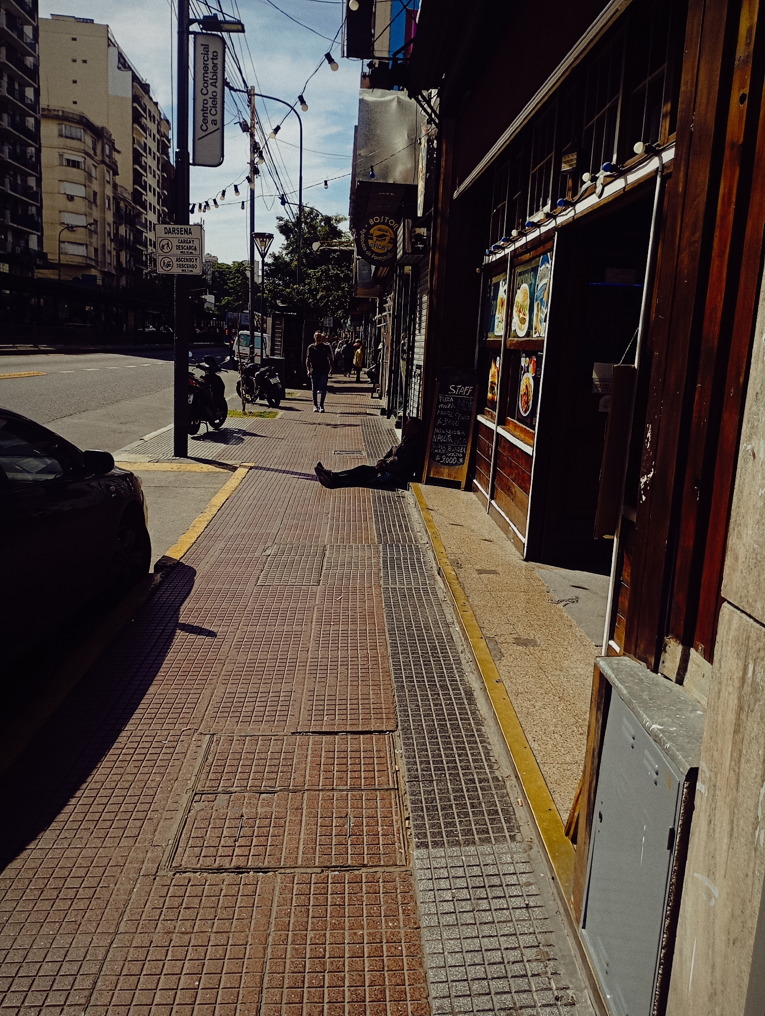 The image depicts a street scene in Buenos Aires, Argentina, on a sunny day. On the left, a street with a black car parked at the curb and a scooter parked further up. Buildings line the street. A tall sign reads "Centro Comercial a Cielo Abierto". A person is walking down the sidewalk. A person is lying on the sidewalk near a closed wooden-front restaurant or bar. A chalkboard menu is displayed outside.