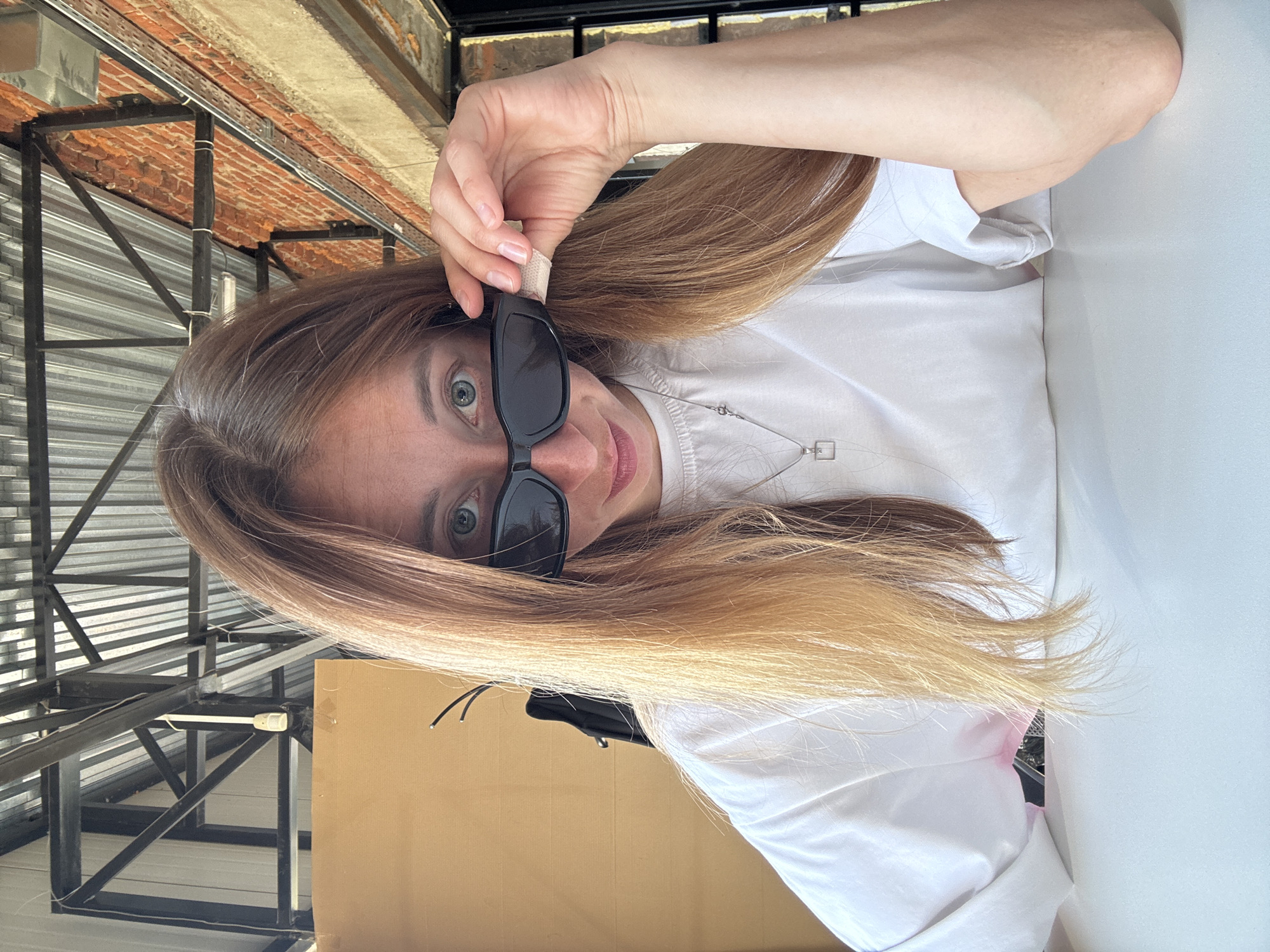 A woman with long, light brown hair is posing for a photo. She is wearing sunglasses and a white shirt. She is holding the sunglasses with one hand. A metal structure is visible in the background. A cardboard box is also in the frame. The location may be Chaikovsky, Russia.