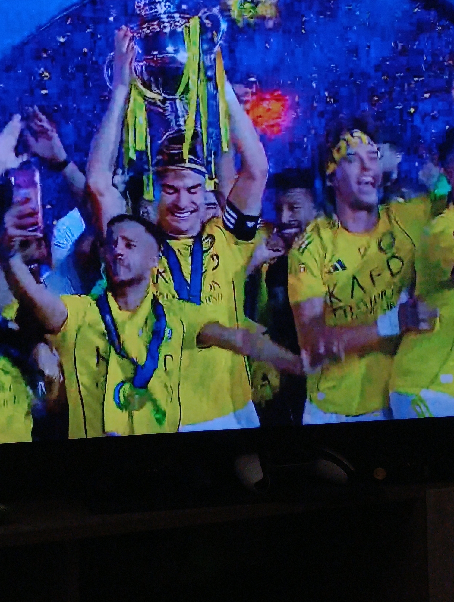 A group of soccer players wearing yellow jerseys and blue medals are celebrating with a trophy. The jerseys have a "KAFD" logo on the front. One player is holding the trophy aloft. Confetti is falling. One player is holding up a mobile phone to take pictures. The event is likely a championship celebration; the location is likely Plovdiv, Bulgaria, based on the team's colors and logos.