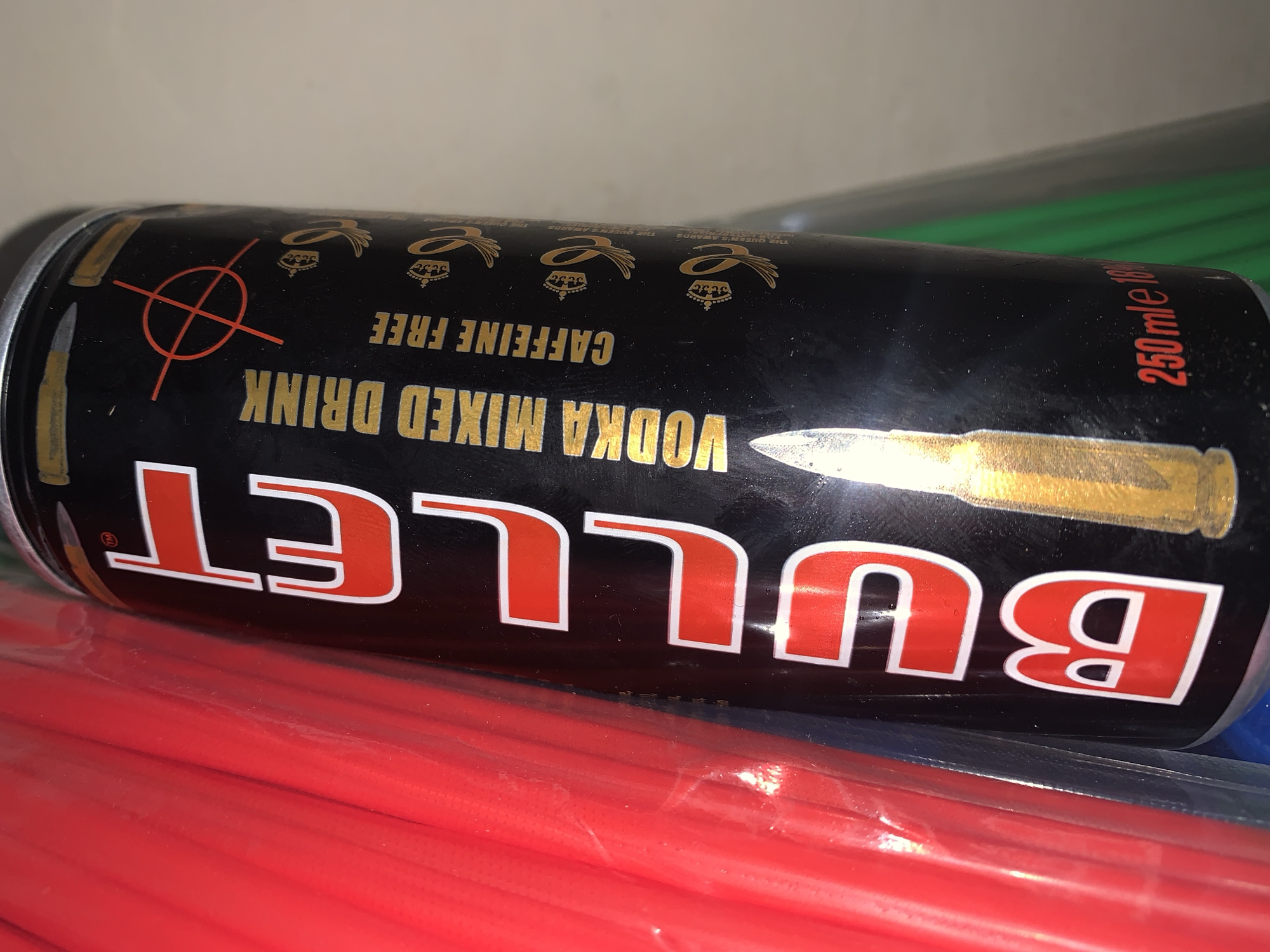 A close-up, eye-level shot shows a black can of "Bullet Vodka Mixed Drink" with gold lettering and graphics, lying horizontally on a pile of bright red cylindrical objects, possibly straws or dowels, enclosed in clear plastic. The can is the primary focus, with its label prominently displayed. The text "BULLET" is emblazoned across the can in large, bold, red letters with white outlines. Above this, smaller text reads "VODKA MIXED DRINK" and "CAFFEINE FREE." There are also several golden crown graphics and what appears to be a target symbol with a red circle and crosshairs, along with a bullet graphic. To the right of the can, the red text "250mle 10%" is visible. The background is out of focus, showing a light-colored wall and a hint of green. The scene is indoors and appears to be a product shot or still life. There are no people or any indication of the city of New Stadium, Nigeria in the image.
