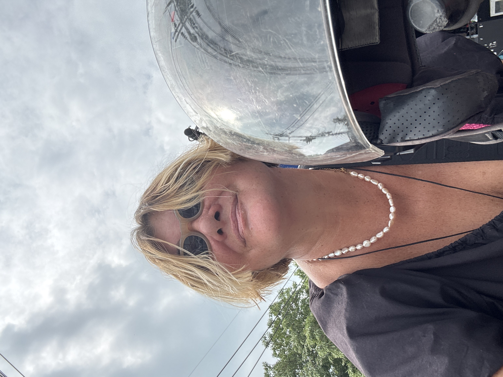 A medium shot from below captures a woman with blond hair, wearing sunglasses and a pearl necklace, seemingly on or near a vehicle with a helmet. The helmet is positioned above her head, partially obscuring the view of the sky, which is overcast. Electrical wires can be seen in the reflection on the visor of the helmet. The woman is wearing a black top. In the background, there is a tree and part of the urban landscape of Baan Plai Laem, Thailand.