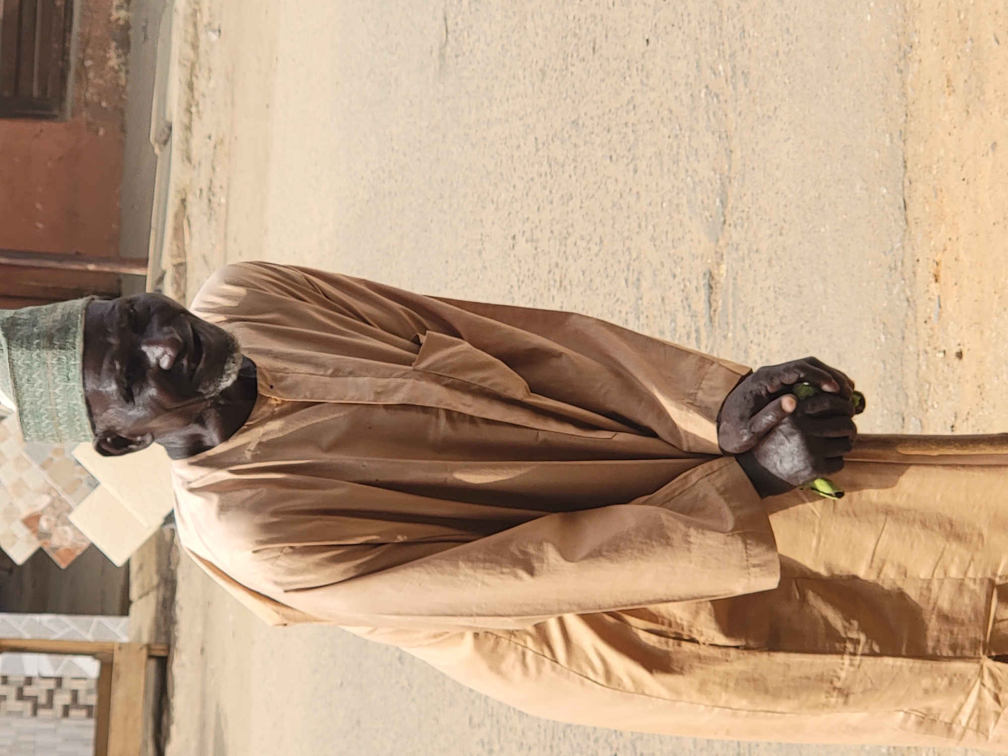 An elderly Black man, facing left, stands in the city of Maiburiji, Nigeria. He wears a long, tan-colored garment and a green cap. His hands are clasped together, holding a light-brown walking stick. He is standing near a building with a partially-visible brown window frame, with white tiles. The background shows a light-brown or beige paved surface, likely a road or pathway.