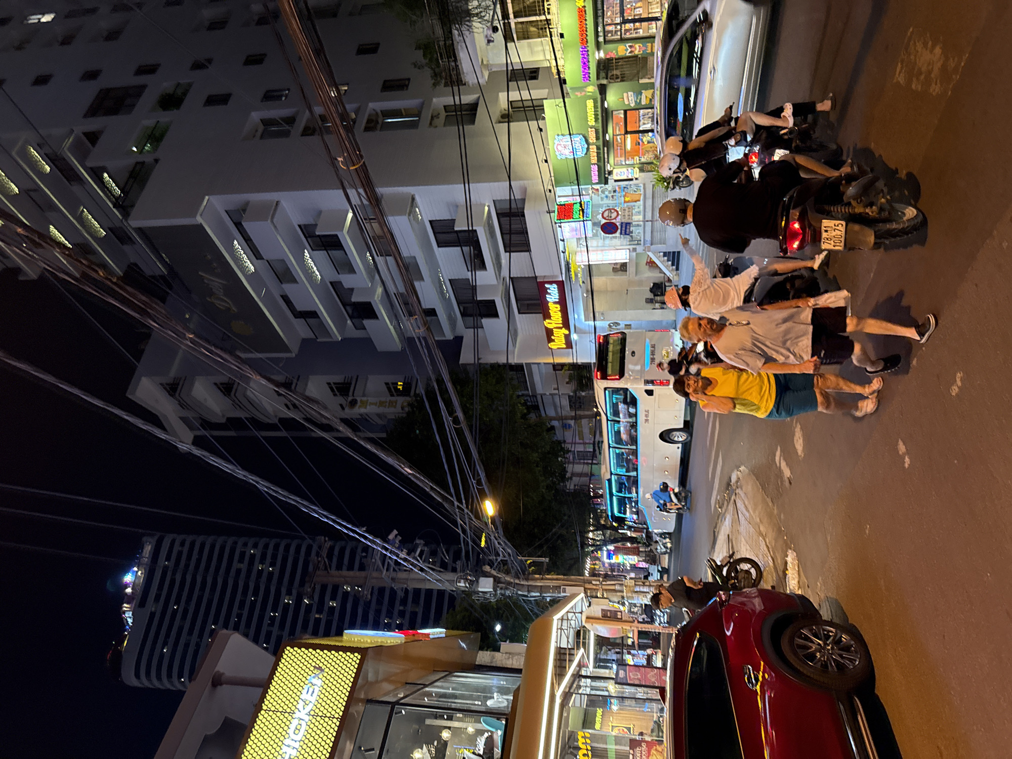 This is a nighttime street scene in Nha Trang, Vietnam, featuring a row of buildings, including the "Daisy Flower Hotel," and several people on foot and on motorcycles. The street is illuminated by streetlights and the neon signs of businesses. A large white tour bus is parked on the right side of the street, and several motorcycles are present, some with riders. A red SUV is parked on the left side of the frame. The architecture suggests a dense urban environment, with power lines crisscrossing overhead. People appear to be casually walking and riding, suggesting a lively commercial or tourist area.