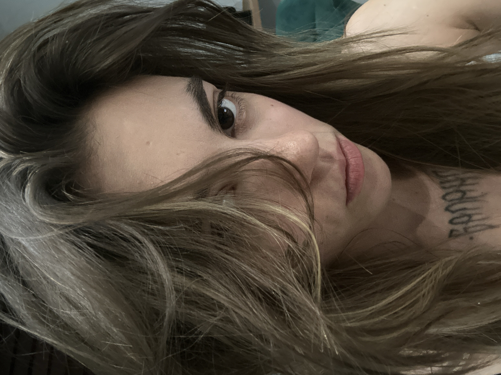The image is a close-up, low-angle, indoor selfie of a young woman with medium-length, layered, brown and blonde hair that partially obscures her face. Her left eye is visible, looking directly at the camera. She has fair skin, and a blurred tattoo is visible on her chest. The background is out of focus, but suggests an indoor setting with a dark wall and a hint of a green object, possibly furniture. There is no indication of the city of Moscow, Russia, or any specific location within it.