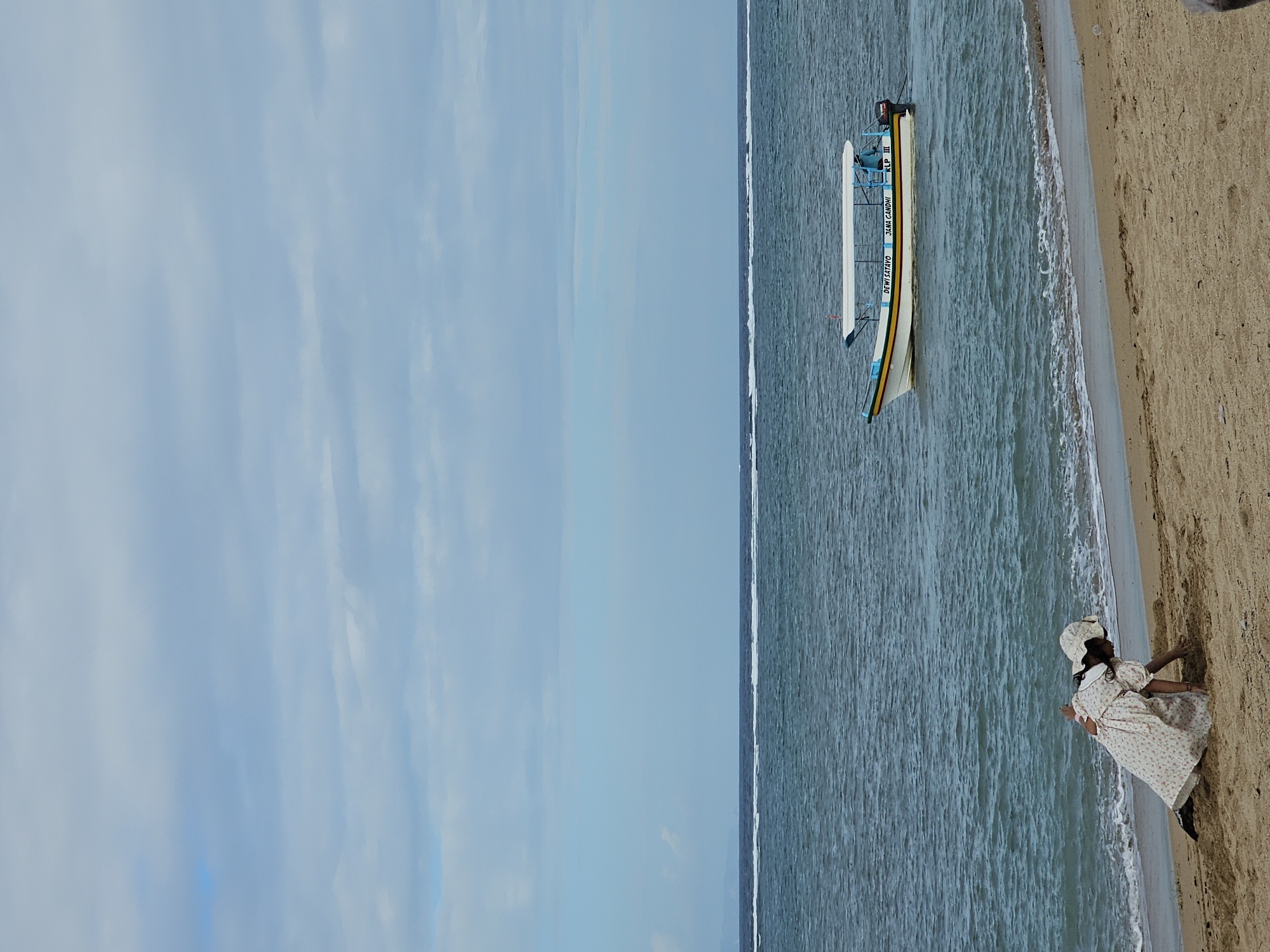 A beach scene in Denpasar, Indonesia, with a child in a dress and hat playing in the sand near the water's edge. A white boat with a blue and yellow stripe is anchored in the blue ocean, with a few whitecaps on the water. The sky is overcast with pale blue and grey clouds. The sand is tan and appears wet near the water.