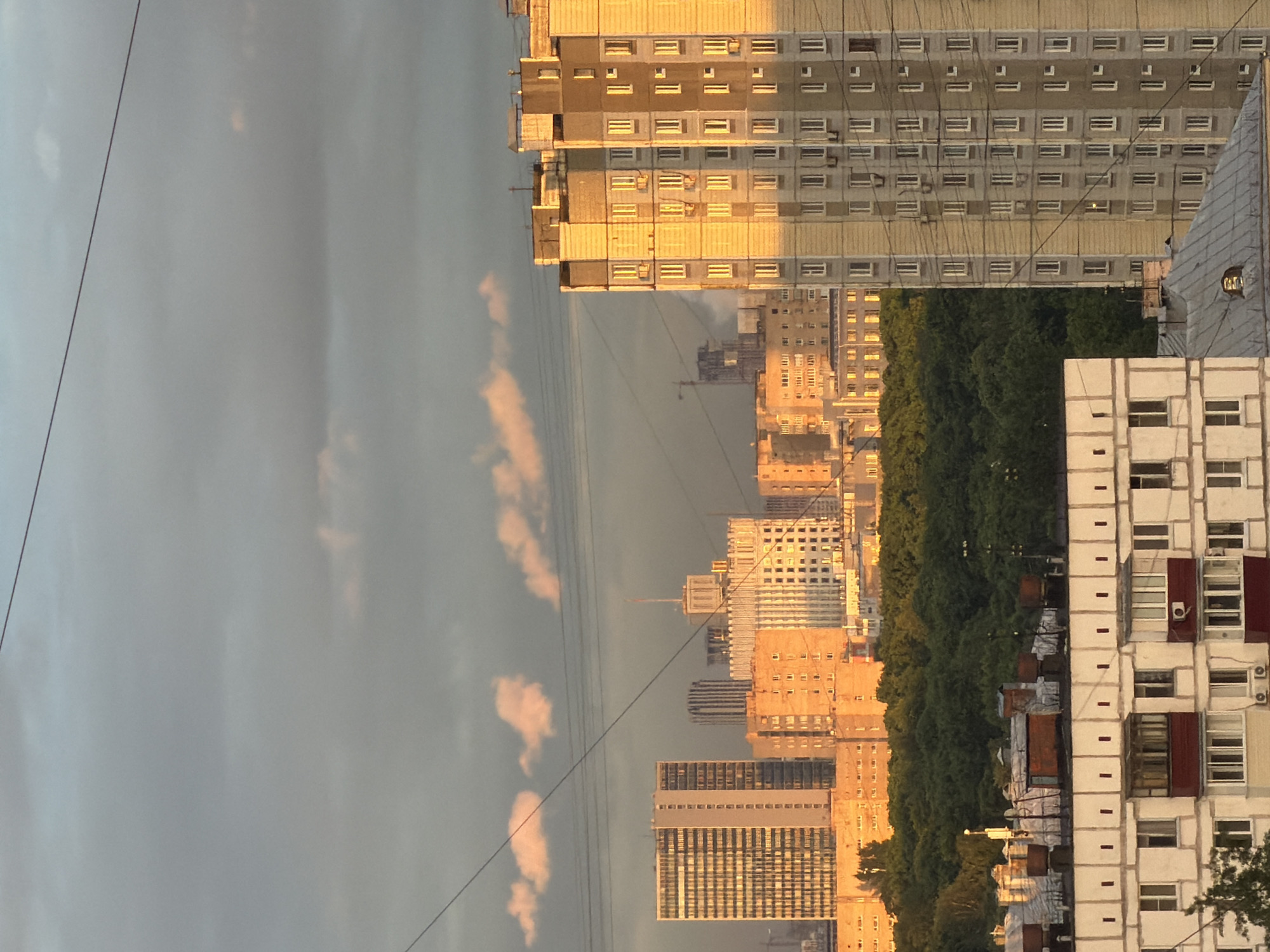 This is an outdoor shot of a cityscape in Khimki, Russia, taken during sunset. Several high-rise apartment buildings, constructed with prefabricated concrete panels, dominate the view. The buildings are arranged in rows along the horizon, separated by a line of trees. The sky is overcast, with clouds appearing as pale pink due to the setting sun. Two large wires run diagonally across the top-left corner of the image.