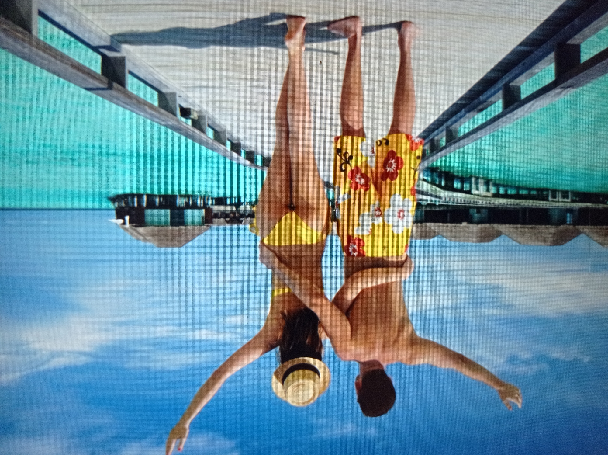 This is a wide-angle, slightly distorted photograph of a couple standing on a wooden pier over turquoise water. The couple is viewed from behind, with their arms around each other. The man is wearing yellow swim trunks with red and white floral patterns. The woman is wearing a yellow bikini and a straw hat. Both have their arms raised in the air, seemingly in celebration. In the background, there are overwater bungalows and a clear blue sky with scattered clouds. The scene appears to be a tropical resort. This image does not depict the city of Mukachevo, Ukraine.