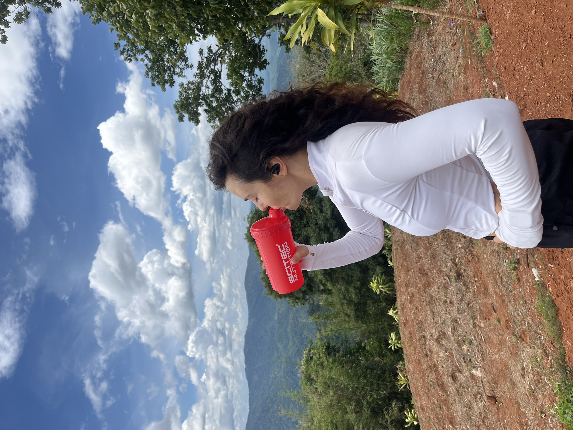 A woman with long, curly dark hair is shown from the mid-torso up, in profile, drinking from a red shaker bottle. She is wearing a white long-sleeved shirt and black pants, and black earbuds are visible in her ears. The background shows a lush, mountainous landscape under a bright blue sky with scattered white clouds. The woman is standing on a reddish-brown dirt slope, with green trees and foliage around her. The location is stated to be Nao Sẻ, Vietnam.