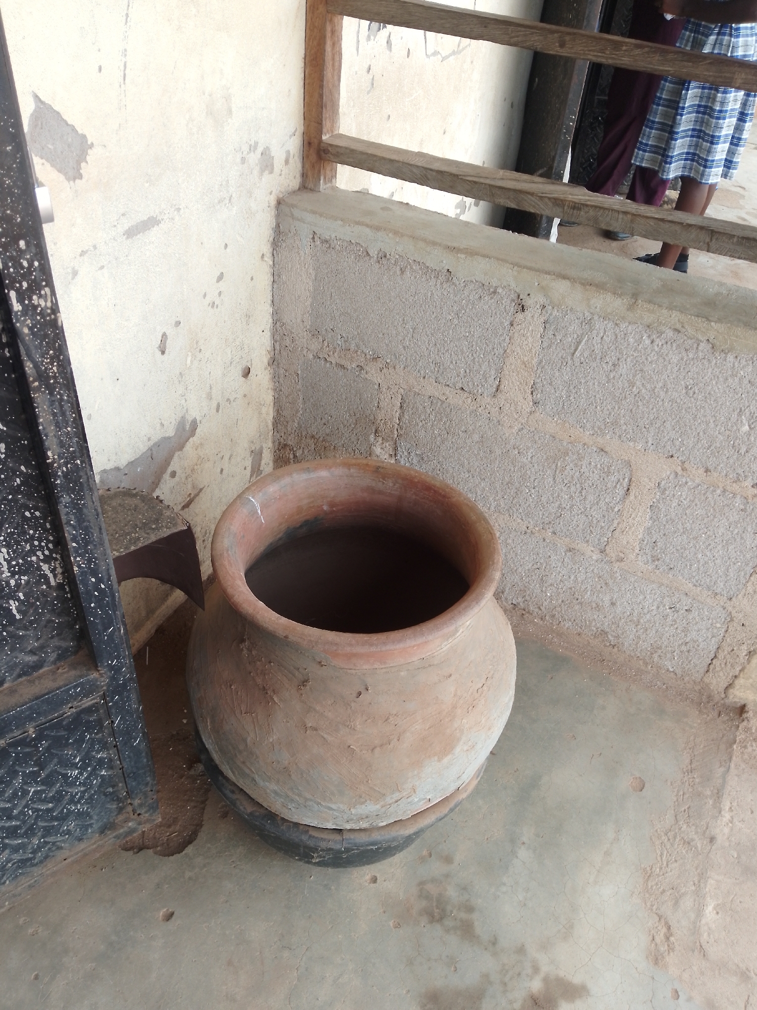 A wide shot captures an outdoor scene in Serti, Nigeria, focusing on a large, tan clay pot. The pot, slightly weathered and resting on a dark, circular base, sits prominently on a light gray concrete floor. To the left, a dark metal gate with white speckles is partially visible, its lower edge suggesting it's either open or its lower section is removed. Behind the pot and to the right, a wall constructed of light brown bricks is visible, with patches of lighter colored plaster. A wooden railing runs horizontally across the upper portion of the image, supported by vertical wooden posts. In the background, beyond the railing, the legs of two individuals are visible; one is wearing a blue and white checkered skirt, and the other is wearing a dark maroon garment and dark shoes. The overall lighting suggests it is daytime, with natural light illuminating the scene.