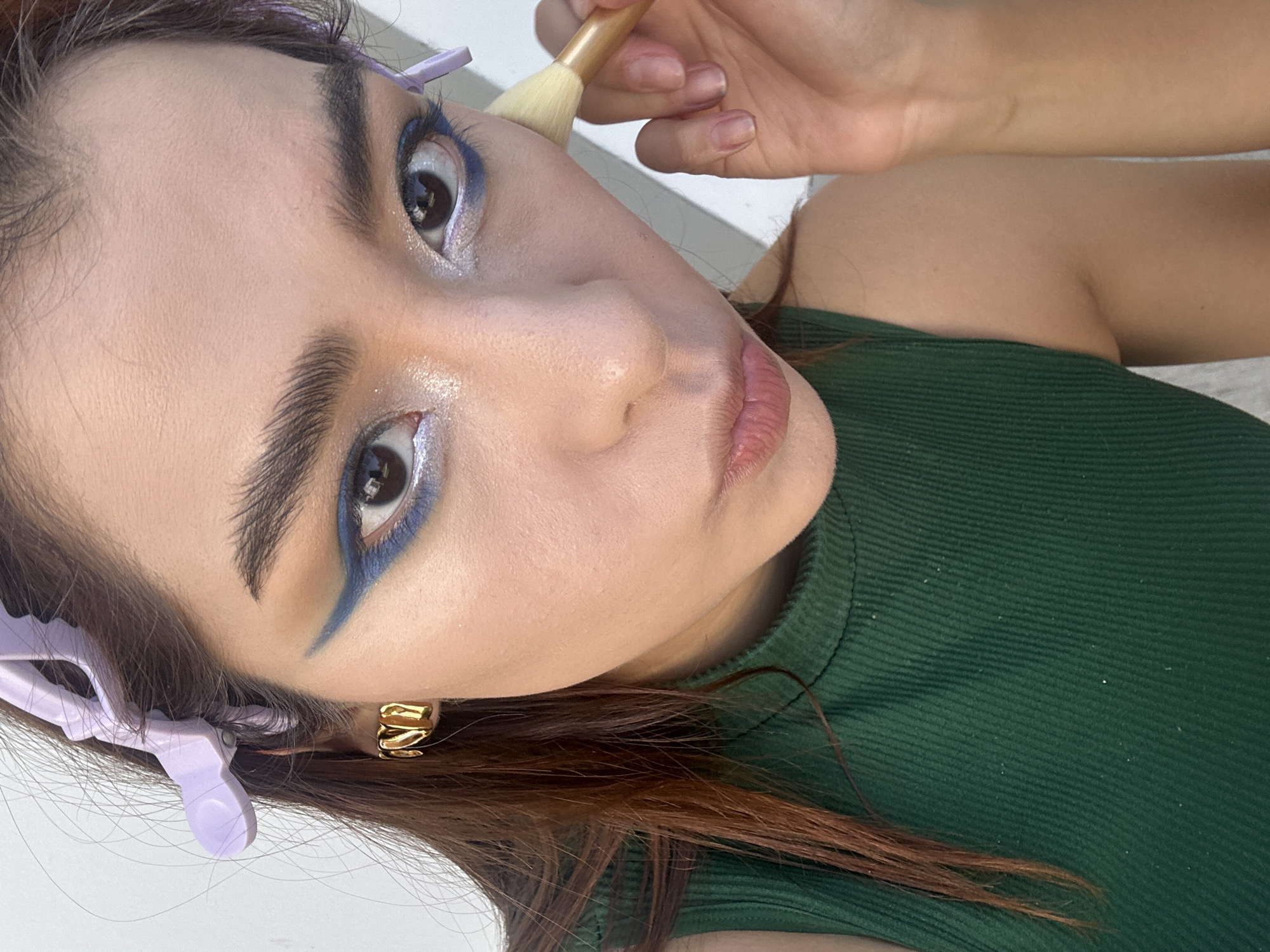 The image presents a close-up, high-angle view of a woman during a makeup application. Her face is the central focus, angled upwards and to the viewer's right. She has pronounced eyebrows and dramatic eye makeup, featuring a sharp, winged blue eyeliner on both upper and lower lash lines, complemented by a shimmering light eyeshadow on her lids and inner corners. Her lips are a subtle pink.
A hand is visible in the upper right, holding a short, light-colored makeup brush against her upper cheek or temple, suggesting the application of powder or highlight. Her dark brown hair is partially pulled back from her face with lavender butterfly clips, one clearly visible near her right temple. She wears a dark green, ribbed top and a gold-toned, multi-band earring on her right ear. The background consists of undifferentiated light-colored surfaces. No text is visible in the image. The activity takes place in San Bernardino Tlaxcalancingo, Mexico.