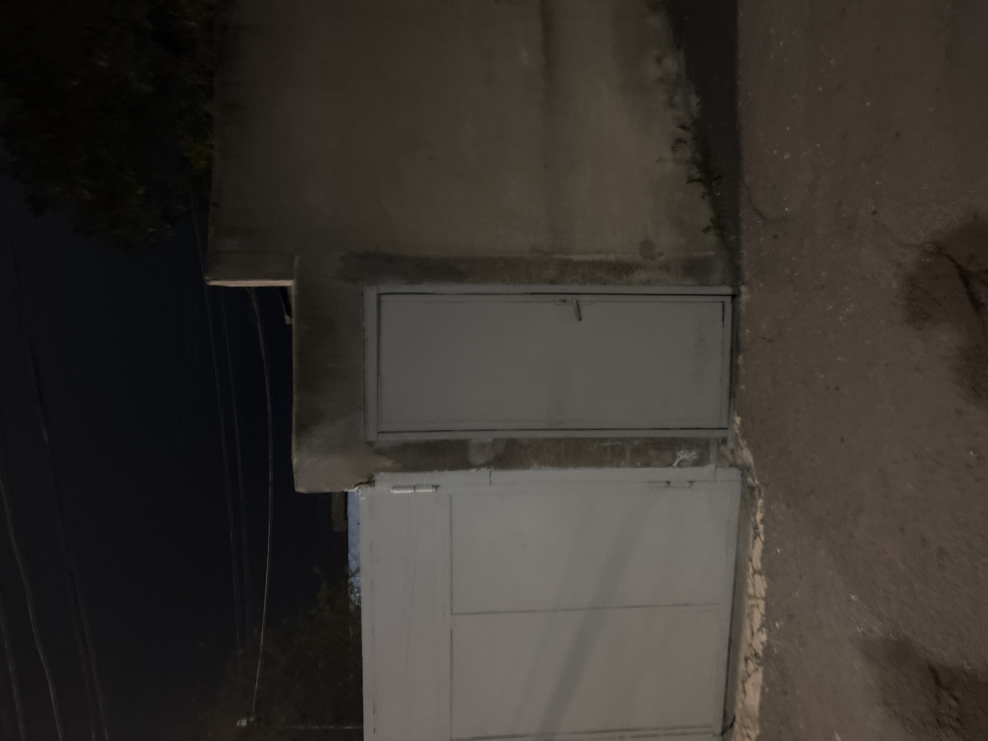 Captured at night, the image shows a section of a building exterior in Yerevan, Armenia. The facade is composed of concrete or a similar material. The structure includes two doors or access panels, one positioned above the other. The top one is smaller and secured with a simple latch, while the lower one is larger with horizontal seams. The surface is slightly weathered. To the left of the building, trees and electrical wires are visible against the dark sky. To the right is a street surface.
