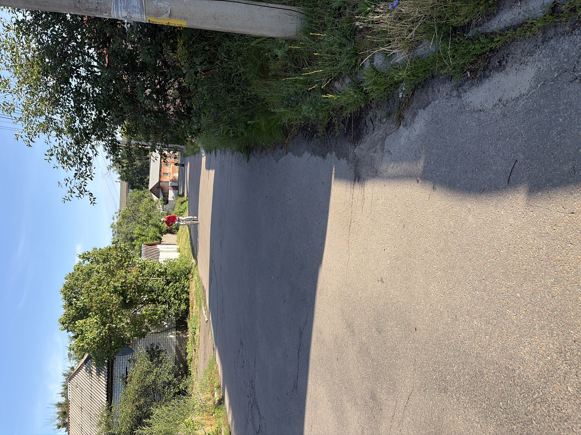 A street scene in a residential area of Chernihiv, Ukraine, under a clear blue sky. An asphalt road dominates the foreground, with a faint shadow cast across its surface from the left. On the right side of the road, a utility pole stands tall, partially obscured by lush green foliage. Further down the road, a person in a red shirt is riding a bicycle, moving away from the camera. Behind the cyclist, a small orange-colored building with a dark roof is visible, and beyond that, trees and other structures. The left side of the road features a white brick building with a grey tiled roof, surrounded by greenery. The overall atmosphere suggests a quiet, sunny day in a somewhat rural part of the city.