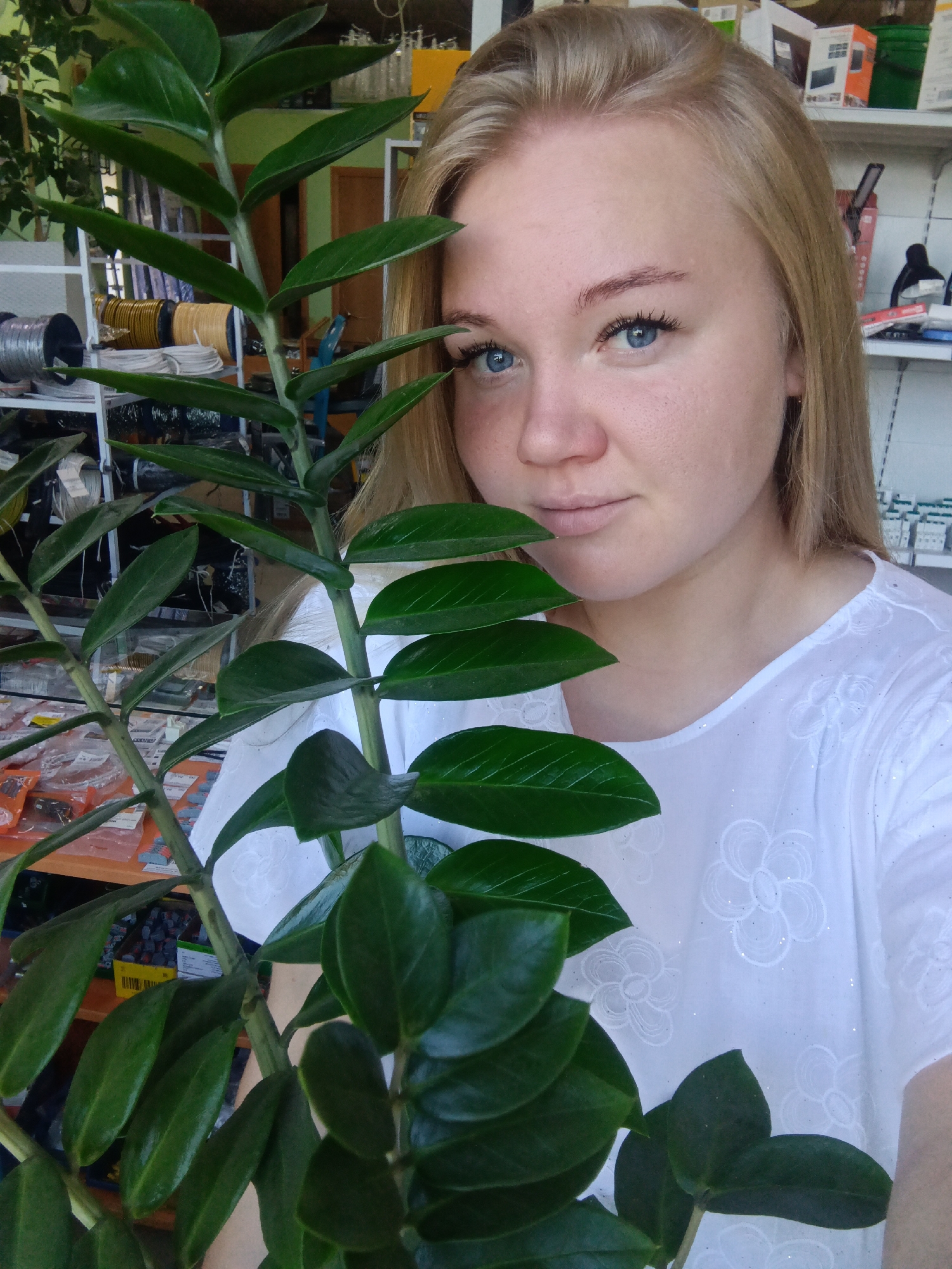 A young woman with blonde hair and blue eyes is holding a large green plant in front of her. She is wearing a white shirt with embroidered flowers. The background appears to be a store or workshop, with shelves filled with various items, including spools of wire and boxes. The lighting suggests an indoor environment. There is no information in the image to determine the specific location in the city of Фоки, Russia.