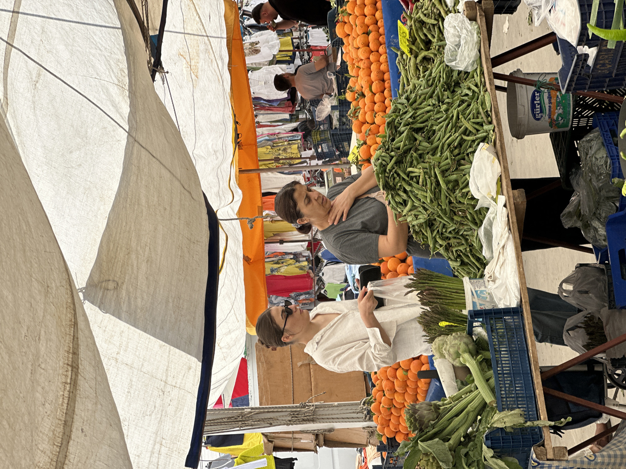 The photograph captures a bustling scene at a marketplace in Fethiye, Turkey. Under a canvas canopy, two women stand at a produce stall. One woman, dressed in a white shirt and sunglasses, inspects a bag, while the other, wearing a gray shirt, looks on. The stall is laden with fresh produce including oranges, green beans, asparagus, and artichokes. Behind the stall, other people mill about, examining clothing. The environment is filled with activity and the typical wares found in a Turkish marketplace.