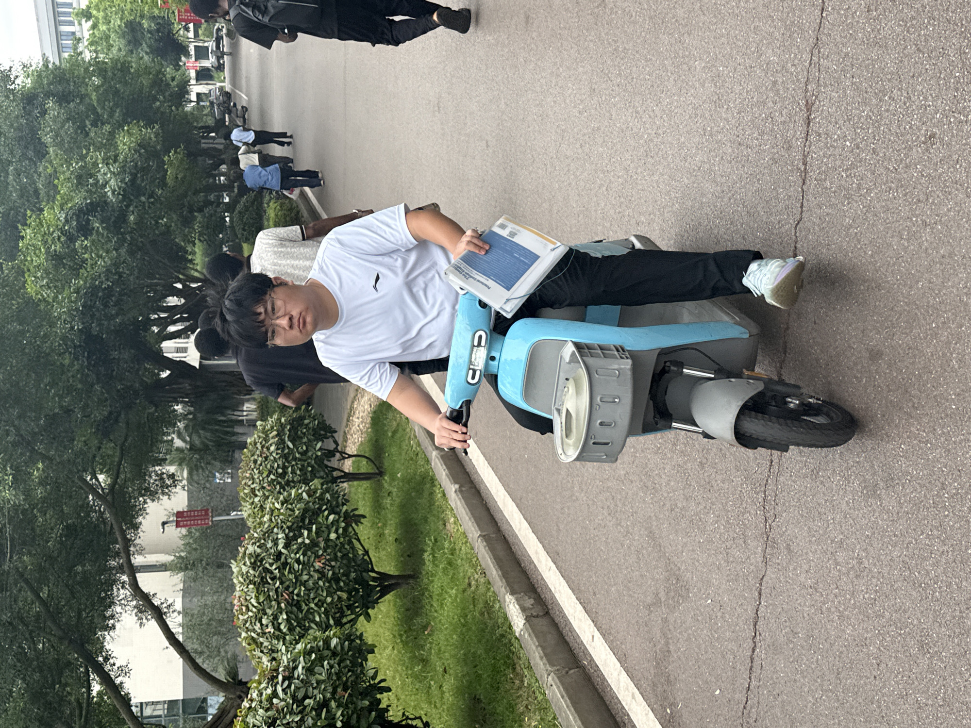 A person wearing glasses and a white t-shirt sits on a light blue scooter. The scooter has a gray basket in front. The person is holding a textbook with a blue cover in their left hand. The person is wearing black pants and white sneakers. The location appears to be a street or pathway near a green area with trees and grass. There are other people walking on the pathway, away from the person on the scooter. Based on the metadata tag provided, the image was taken in the city of Jiangjin, China.
