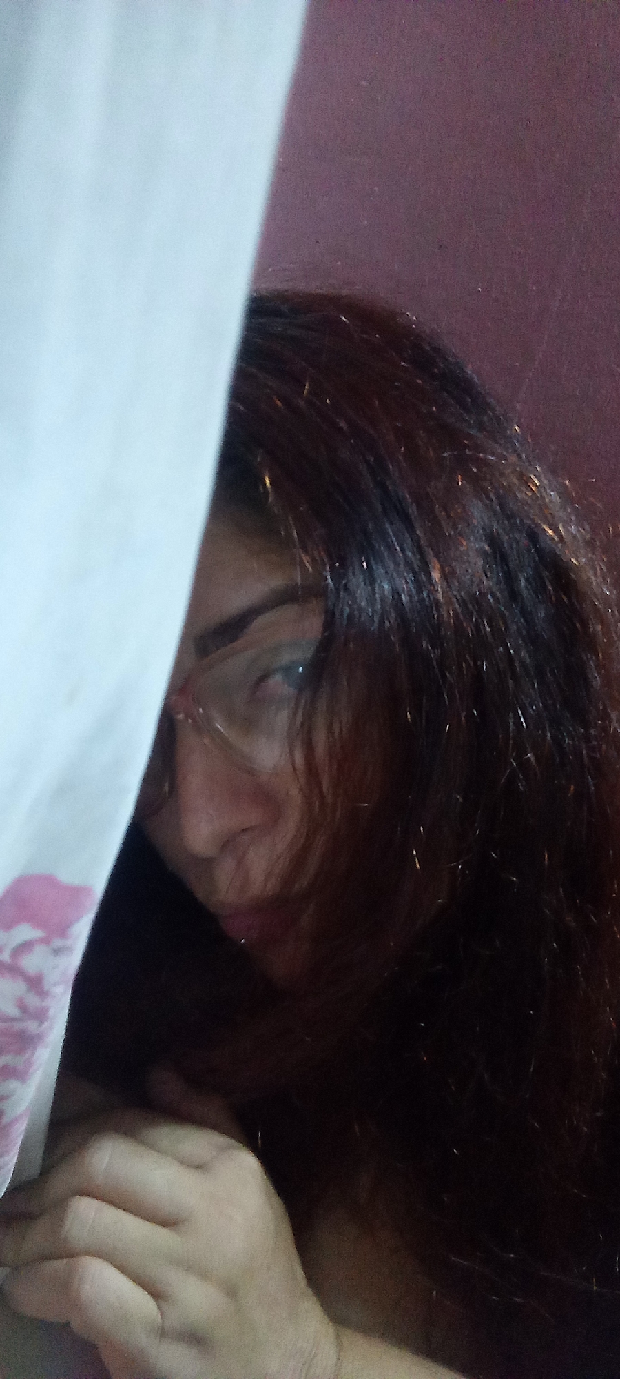 A woman with reddish-brown hair and glasses partially obscured by a white curtain is visible. Her face is slightly tilted, and her hand is grasping the curtain. The background appears to be a plain, light-colored wall. The location is unknown.