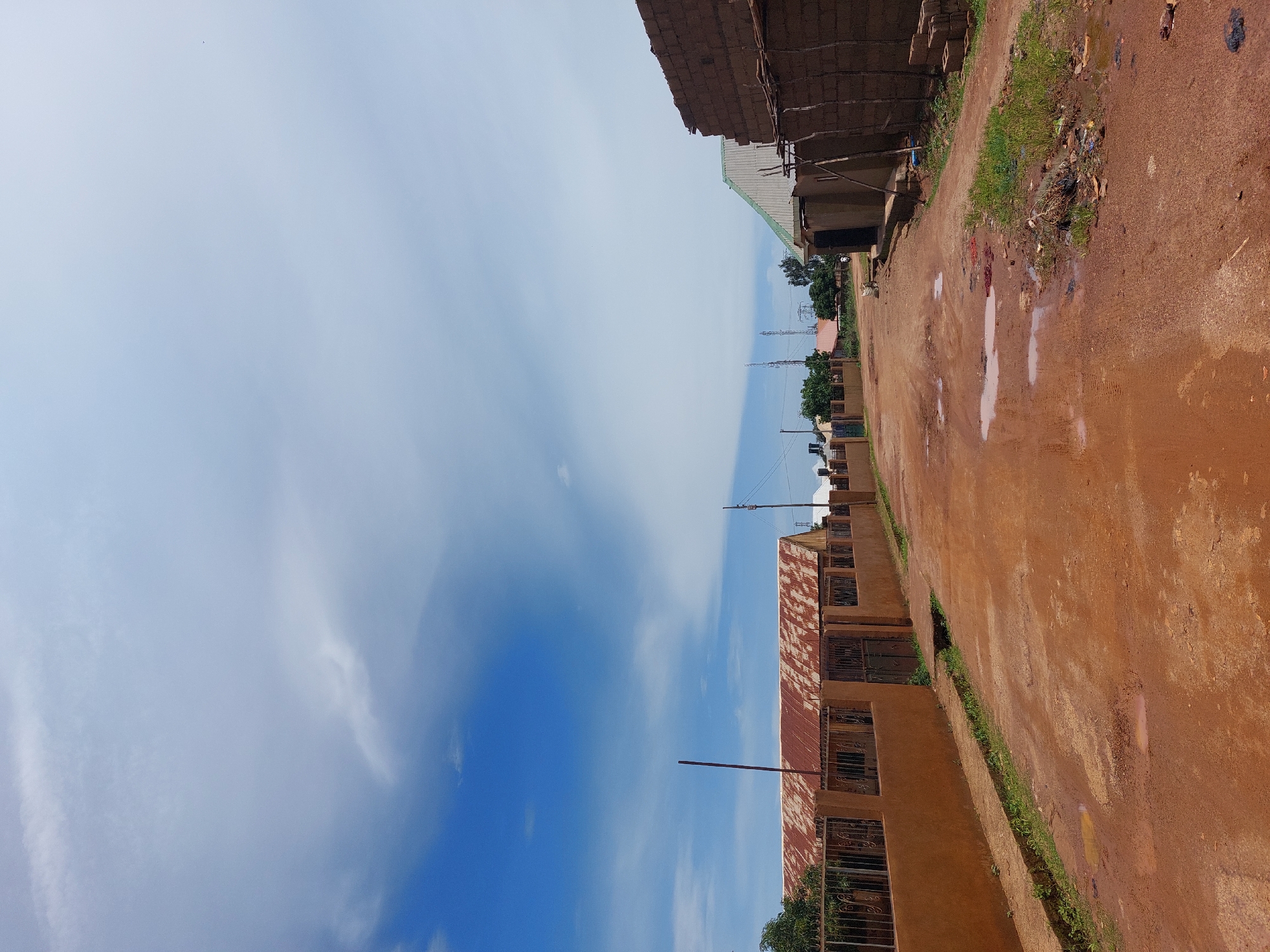 The image depicts a dirt road in Barakin, Nigeria, under a partly cloudy sky. The road is reddish-brown and appears to be in a rural or semi-urban area, with puddles of water indicating recent rainfall. On the left side of the road, there is a long, tan-colored wall topped with a rusted corrugated metal fence, behind which are buildings with similarly rusted roofs. A tall, thin pole stands in front of this wall. Further down the road, trees and some vegetation are visible. On the right side of the road, there are unfinished buildings constructed with mud bricks and some wooden supports. Scattered debris and vegetation can be seen along the roadside, particularly on the right. The overall impression is of a developing or modest community, with a focus on the unpaved infrastructure and basic construction.