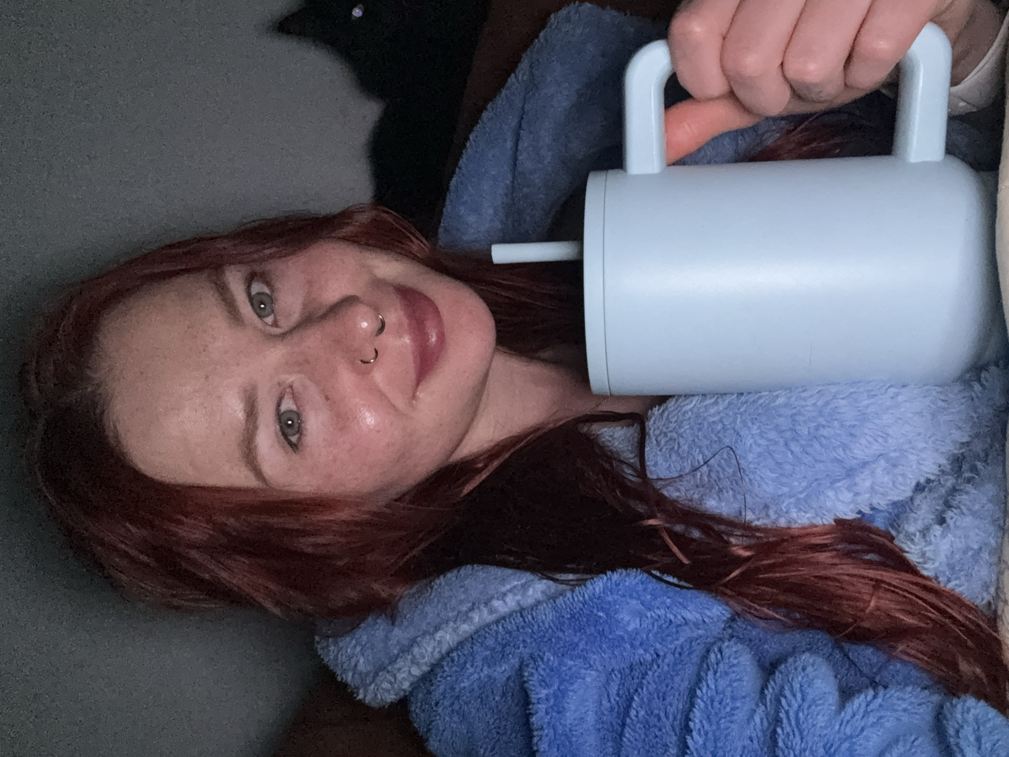 A woman with red hair, a nose ring, and visible freckles is lying down while holding a light blue drinking mug with a handle and straw. She is wearing a blue fuzzy robe. The background includes a dark area, possibly a wall or another object. This image does not provide any information regarding the location in the city of Rostov-on-Don, Russia.