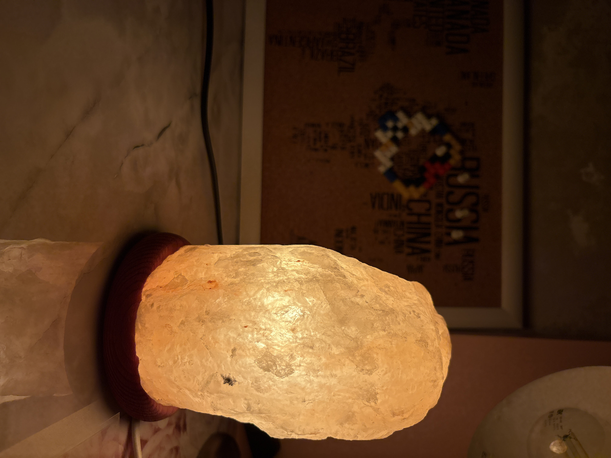 This media file captures a close-up of a Himalayan salt lamp, casting a warm, orange glow. The lamp itself is irregular in shape, resembling a rough-hewn crystal. It is supported by a dark red, ribbed base.
In the background, a corkboard is visible, adorned with what appears to be a world map made of small, colorful squares or beads. Text is also visible on the corkboard, including country names like "RUSSIA," "CANADA," "INDIA," and "CHINA." The text is printed in dark letters against the brown cork material.
The scene is dimly lit, suggesting it might be nighttime or an interior setting with ambient lighting. The overall mood is cozy and relaxed, illuminated by the soft light of the salt lamp. The setting is likely a home or a personal space, given the decorative elements. There are no people visible in the frame. The location context provided is Засечное, Russia, which may explain the presence of text related to Russia on the corkboard.