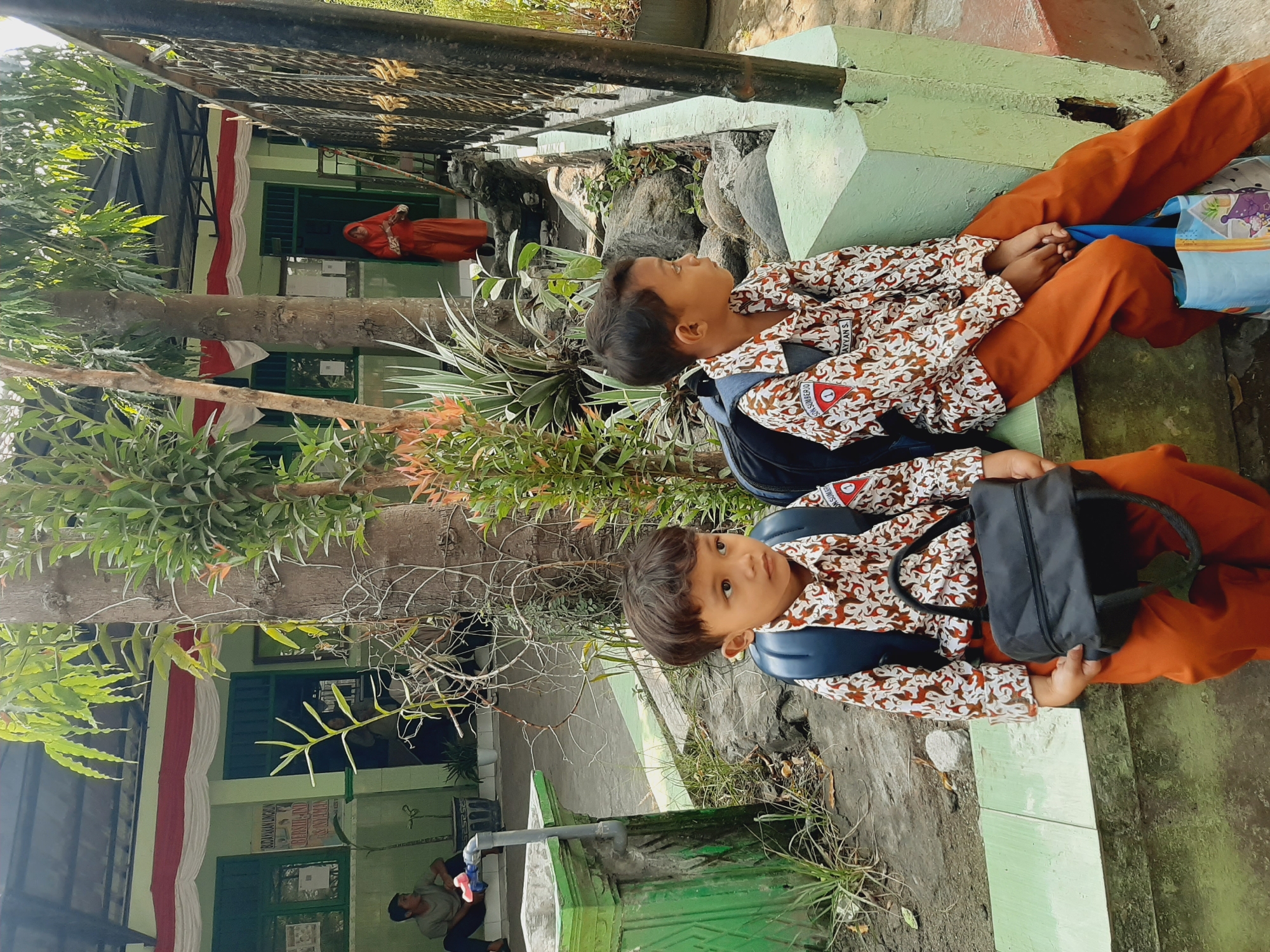 In a school setting in Sumberjo, Indonesia, two young boys, likely students, sit on a low green wall. They are dressed in white shirts with a brown and white pattern and orange pants, typical school uniforms. Both boys wear backpacks. The boy on the left holds a black bag on his lap, while the boy on the right has a blue shopping bag near his feet.

The school building behind them is painted green with a red and white draped banner along the top of the facade. Several windows are visible. A person in an orange hijab stands in the doorway of the building. To the left of the building, a young man in a grey t-shirt and dark pants sits on steps.

The environment is outdoors with a paved walkway and some rocky terrain. Trees and various plants, including spiky green ones, surround the area. A metal fence is visible on the right side of the frame. The lighting suggests it is daytime, with sunlight casting shadows.