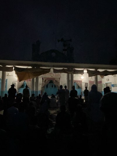A large group of people are gathered outdoors, seemingly in prayer, under a covered area with columns. The scene appears to be at night or in very low light. In the background, a building with towers is visible. The location is most likely Kandahar, Afghanistan.
