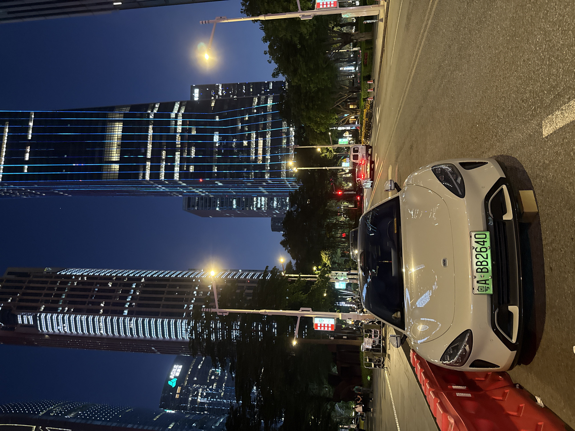 The image depicts an urban scene at night in Tianhe District, Guangzhou, China. Dominating the background are illuminated skyscrapers, one featuring horizontal blue light lines. A white electric car, identified by the 'MI' logo on its front, is parked in the foreground against a red barrier. The car has a green license plate marked "粤A·BB2640". The street is paved and lined with trees and streetlights. Other vehicles are present in the distance. The environment is well-lit, reflecting the vibrancy of the urban district.