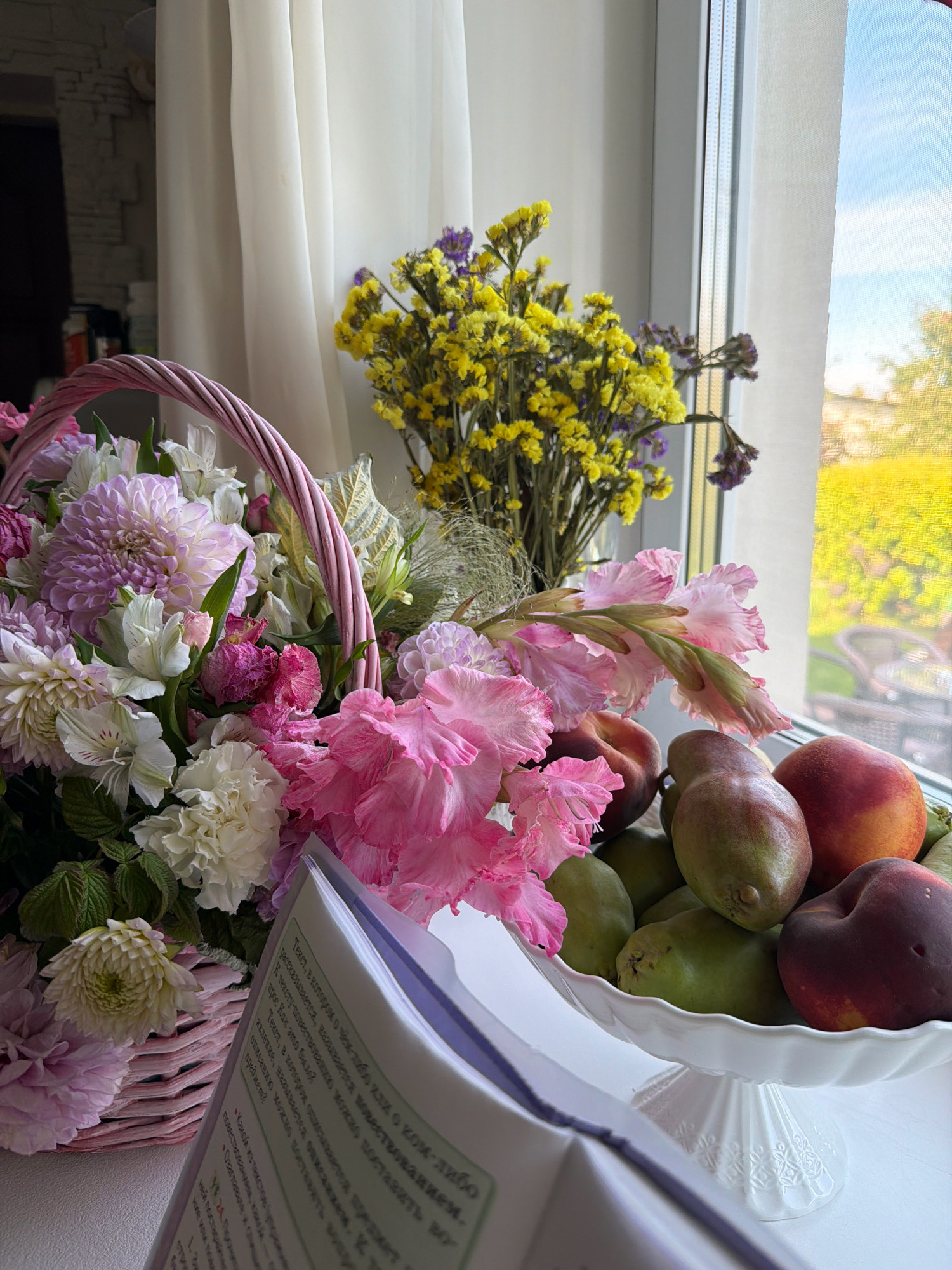 The image shows a still life composition on a windowsill. In the foreground, a book with Russian text is partially open. To the right of the book, a white ceramic cake stand holds a selection of fruits, including pears and peaches. Behind the fruit, a bouquet of pink gladiolus flowers is prominently featured. Further back and to the left, a woven pink basket overflows with a variety of flowers, including dahlias and other white and pink blooms. Behind the flowers, a bundle of dried yellow and purple flowers stands near a sheer white curtain. The windowsill overlooks a bright, sunlit outdoor scene with trees and some buildings. The overall impression is one of a domestic setting, likely a home in Бабініцкі сельскі Савет, Belarus, given the context. No people are visible in the image.