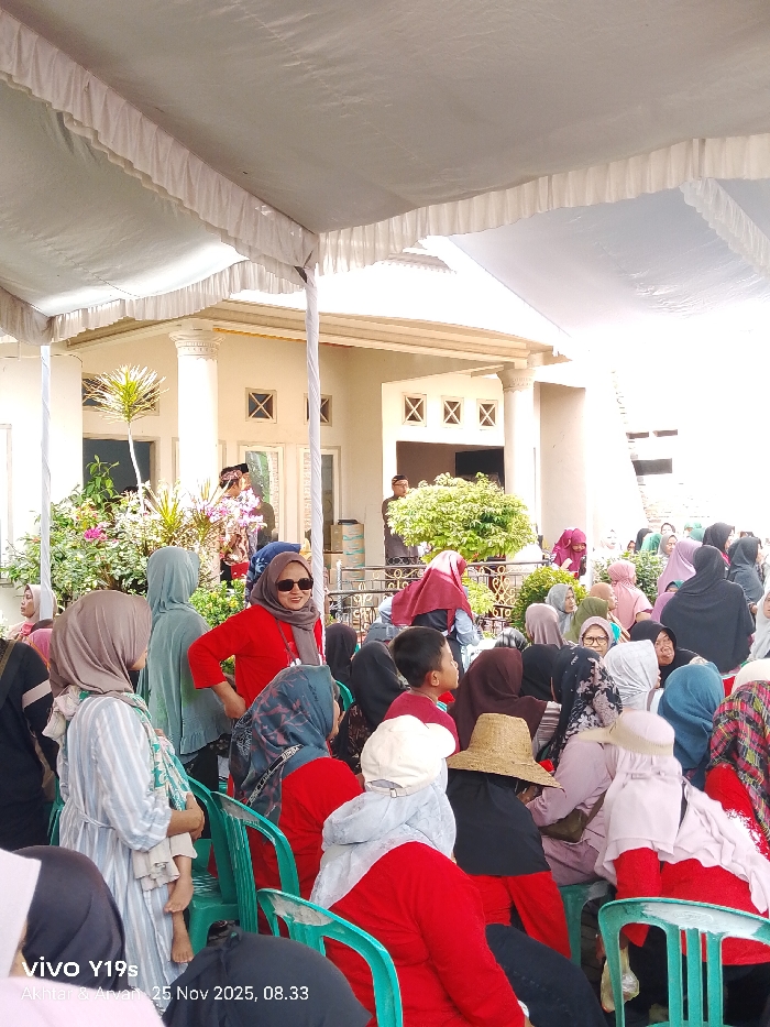 Under a white tent, a crowd of people, mostly women wearing hijabs in various colors and patterns, are seated on green plastic chairs. One woman in a red top and sunglasses stands out. In the background, a building with decorative windows and pillars is visible amidst lush greenery. The scene appears to be an outdoor event or gathering.