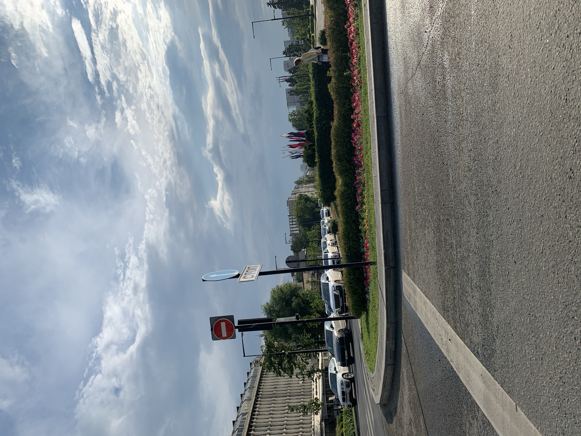 This image depicts a wide, asphalt road in a city setting, likely Perm, Russia, based on the traffic signs. In the foreground, a white line marks a crosswalk or lane divider. The road curves to the left, leading to a roundabout with a circular flower bed. The flower bed is planted with numerous pink and red flowers, bordered by green shrubs and a gray concrete curb.
Beyond the roundabout, traffic is visible, including several cars such as a white SUV and a dark-colored sedan. To the left, a large, multi-story building with a facade of vertical concrete elements dominates the skyline. Adjacent to the building are several trees.
Traffic signs are prominently displayed on poles. One sign is circular with a red border and a white horizontal bar, indicating "No Entry" or "Do Not Enter." Another sign is blue with a white arrow pointing to the right, signifying a mandatory turn. A third sign, also on a pole, is rectangular with white text on a blue background, reading "СТОП" (STOP in Russian).
Further in the background, more buildings and trees are visible. A line of flags, some appearing to be national flags, is planted in a grassy area. A lone individual, a woman with light-colored hair wearing a yellow jacket and blue shorts, walks past the flower bed on the right side of the image. The sky is partly cloudy with dramatic, textured clouds. The overall scene suggests an urban environment on a bright, possibly overcast, day.