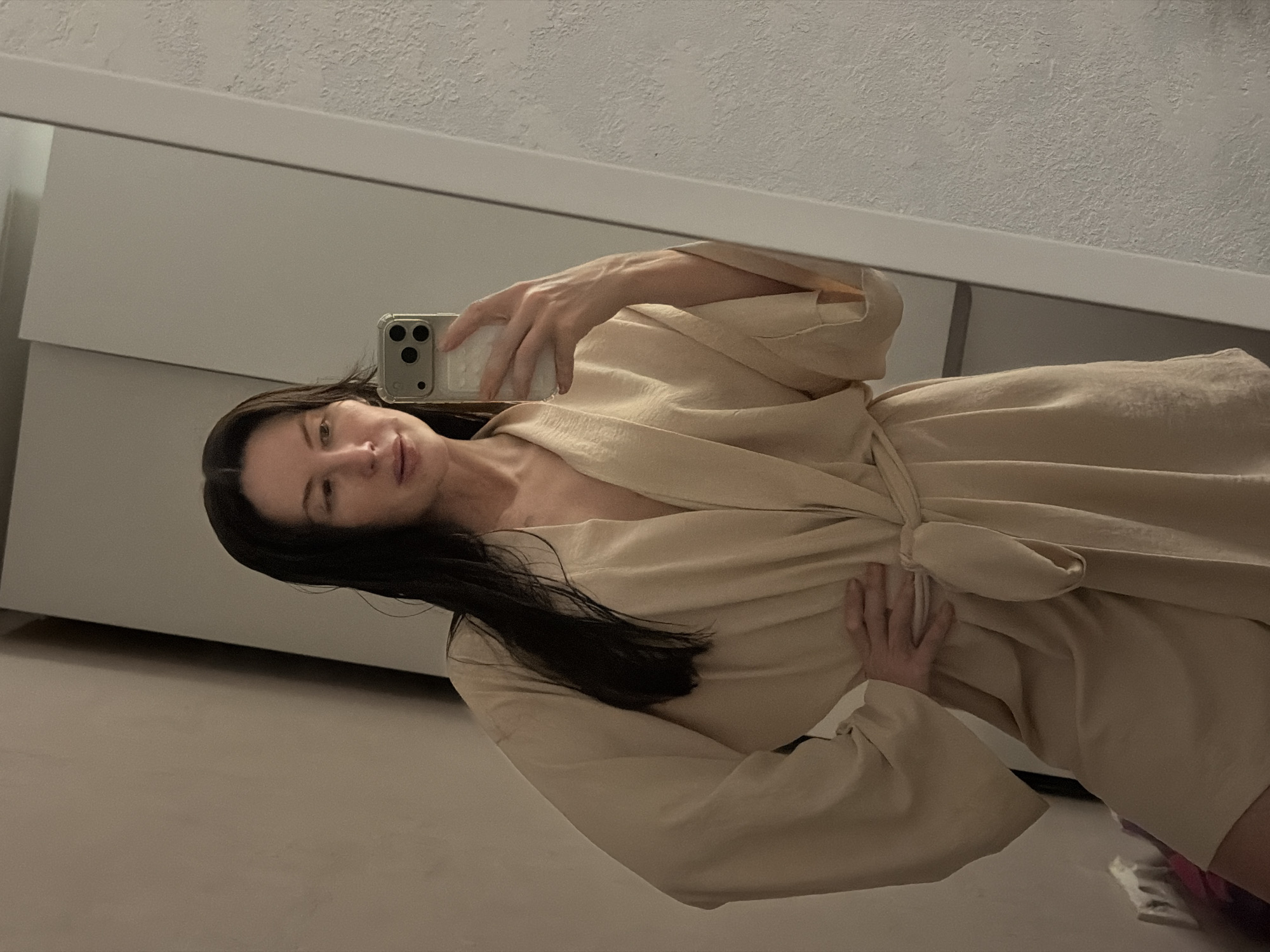 A woman is taking a selfie in front of a mirror. She is wearing a light-colored robe. The background includes a wall and what appears to be a dresser or cabinet. There is no indication of the city of Perm, Russia in the image.
