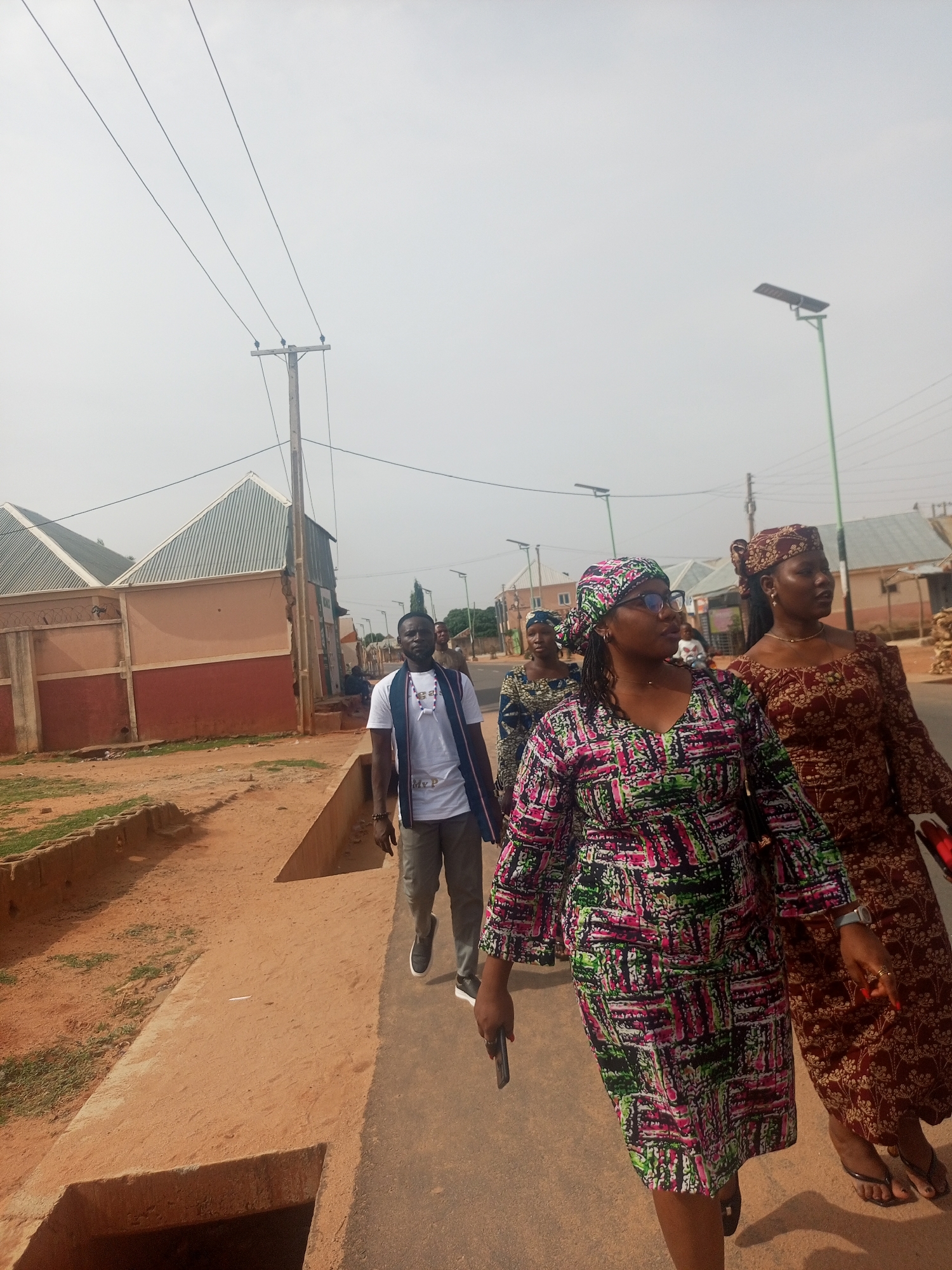 A group of four people are walking along a sidewalk next to a street in a residential area. The person closest to the camera is a woman wearing a colorful patterned dress and headscarf, and glasses. Behind her is another woman in a brown patterned dress and headscarf. Following them is a third woman wearing a similar headscarf and dress, partially obscured by the others. At the back of the group is a man wearing a white t-shirt, grey pants, and a blue scarf around his neck.
The sidewalk is concrete and borders a dirt area with a drainage ditch. Buildings with corrugated metal roofs and red-and-white painted walls are visible in the background. Overhead power lines and a solar-powered street lamp are also present. The sky is overcast.
The location is Tempure, Nigeria.