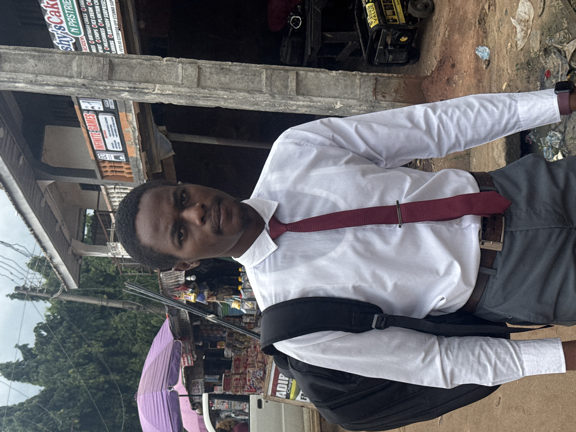 A young Black man stands in front of a street-level business in Ihiagwa, Nigeria. He wears a white collared shirt, a maroon tie, and gray trousers, with a black backpack slung over his left shoulder. In the background, a multi-story building houses businesses, including "Shy's Cake & Pastries" and "INFINITE GAMES". The street is lined with various stalls and structures, some selling goods like drinks and what appear to be automotive fluids. A generator is visible to the right of the man. The environment suggests a bustling urban or semi-urban commercial area.