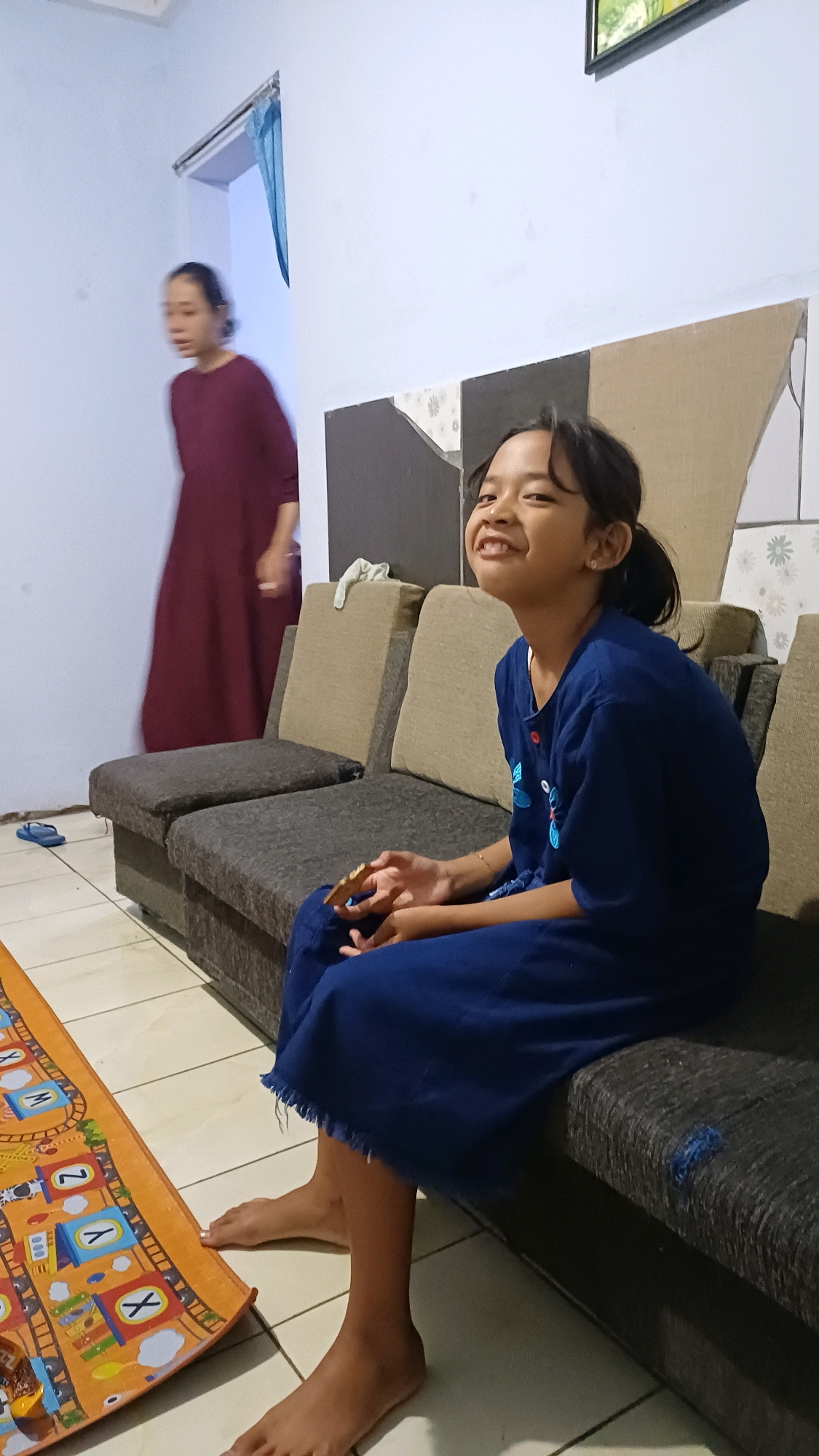 In the living room of a home in Kowangan, Indonesia, a young girl with dark hair pulled back in a ponytail is seated on a dark gray sofa, wearing a blue dress. She is smiling and looking up and to her right, holding a piece of food in her hands. To her left, another person in a dark red dress is walking away, out of focus. A colorful play mat with letters and images of trains is visible on the tiled floor in the foreground. The wall behind the sofa has a decorative panel with a pattern of alternating brown and light gray squares, and a framed picture hangs on the wall above. A pair of blue flip-flops are on the floor near the sofa.