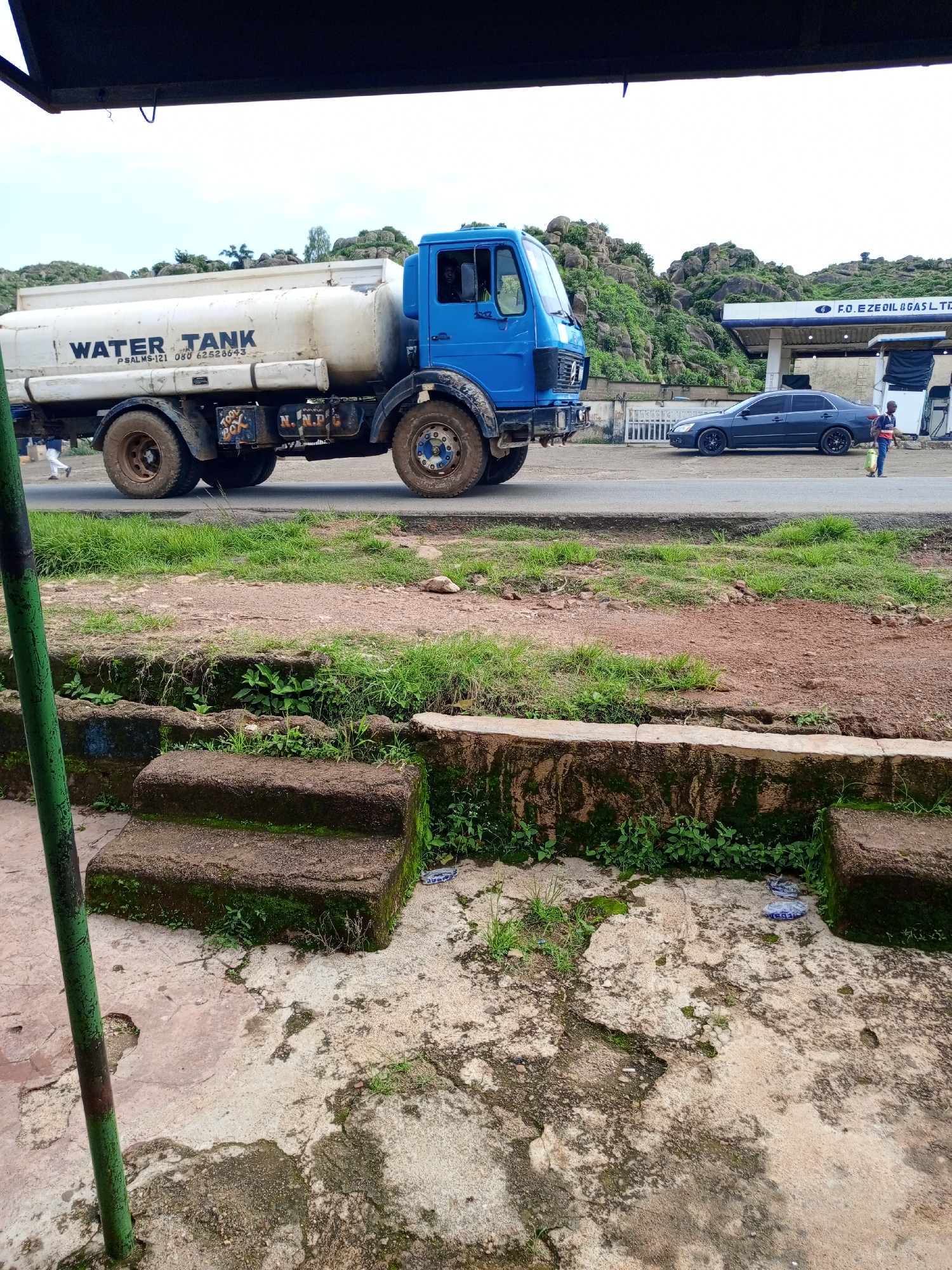 A blue and white water tank truck is parked on the side of a road. The truck has "WATER TANK" written on the side of the tank. Behind the truck, there is a gas station with pumps and a sign that reads "F.O. EZE OIL & GAS LTD". There is a dark gray car parked in front of the gas station. A person is walking away from the gas station carrying a yellow container. In the foreground, there are steps leading up to a moss-covered surface. The road is lined with grass and dirt. In the background, there are rocky hills covered in green vegetation. The sky is overcast. The scene appears to be in Rukuba Road, Nigeria.