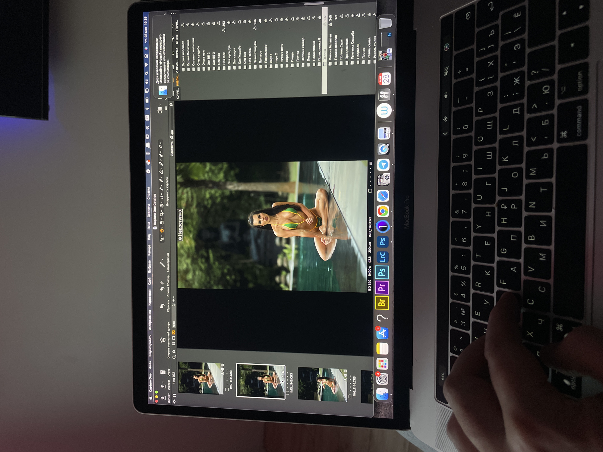 On a table, there's an open MacBook Pro displaying the Capture One software, a photo editing application. The software shows a semi-nude woman posed in a pool of water against a leafy background. Multiple similar images of the woman are displayed below the main image. The left-hand side of the screen features folder listings, and the right-hand side shows editing details, including file names and settings. A hand is visible on the right-hand side of the keyboard, likely interacting with it.
The keyboard itself is set to Cyrillic layout, indicating it may be from a Russian-speaking region.
The sidebar on the left of the main image includes folders such as:
- "Оксана Аэропорт" (Oksana Airport)
- "Оксана Касаткина" (Oksana Kasatkina)
- "Оксана с малышом" (Oksana with baby)
- "Олег свадьба" (Oleg wedding)
- "Ольга муж" (Olga husband)
- "Оля ВБ 2" (Olya VB 2)
- "Оля ВБ 3" (Olya VB 3)
- "Оля Кзн" (Olya Kzn)
"Kzn" would indicate the city of Kazan, Russia.