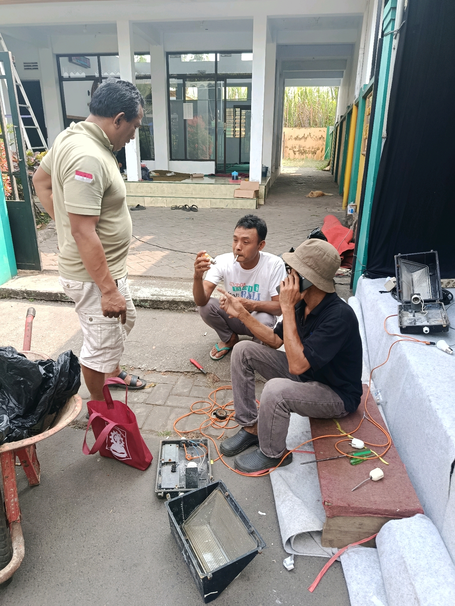 In Bululawang, Indonesia, three men are gathered outdoors near a building. The man on the left, wearing a light olive green polo shirt and light-colored cargo shorts, stands and talks with the other two. The middle man, in a white t-shirt, crouches while smoking a cigarette and holding a small, light-colored object, possibly food, in his hand. The man on the right, wearing a dark shirt and a tan hat, also crouches and speaks on a black mobile phone.
The ground is paved with interlocking bricks, and several electrical components are scattered around, including two open floodlights, orange extension cords, and a light bulb. A red bag and a black trash bag in a wheelbarrow are also visible. In the background, a tan dog lies on the ground in front of a building with white pillars and glass doors. The scene suggests a casual outdoor gathering or a break during a task involving electrical equipment.