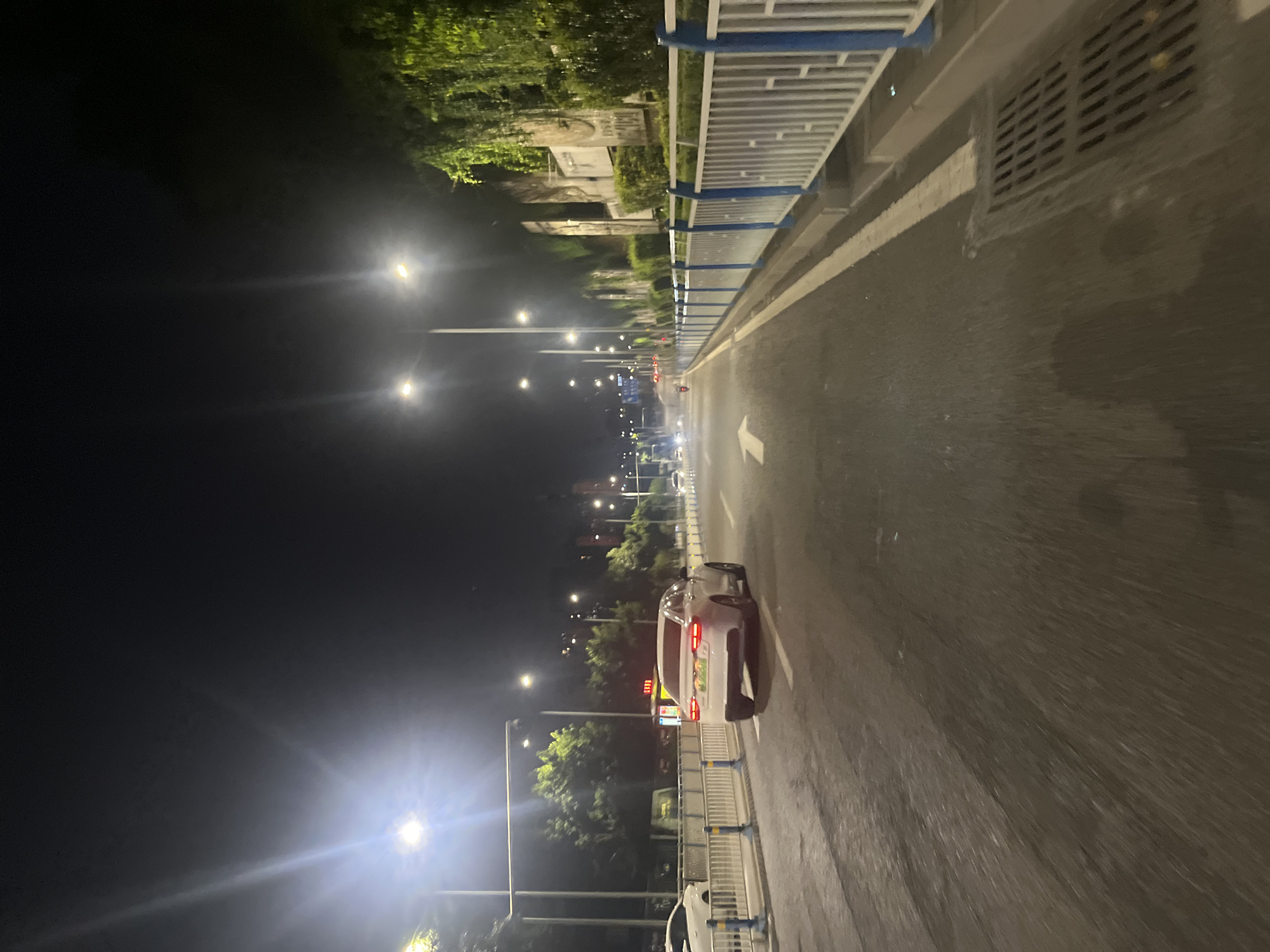 This nighttime photograph captures an urban street scene in Jiulongpo District, China. The asphalt road, appearing damp and reflecting light, extends forward into the distance. A large white arrow painted on the right lane indicates a straight-ahead direction. A white four-door sedan, equipped with a green license plate displaying "A S26516," is observed in the left lane, moving away from the viewer, with its red taillights illuminated. Another white sedan is partially visible on the far left, positioned behind a blue and white metal barrier. Numerous bright streetlights line both sides of the thoroughfare, casting strong illumination and causing noticeable lens flare in the upper left of the image. Blue and white metal barriers delineate the edges of the road on both the left and right sides. Behind the right barrier, extensive green vegetation, including vines and bushes, covers a sloped wall or embankment, with several large, light-colored rectangular panels or signs partially visible within the foliage. On the left, beyond the barriers, additional green trees and structures featuring illuminated reddish-orange elements are discernible. A storm drain grate is situated at the far right edge of the roadway. No individuals are visible.