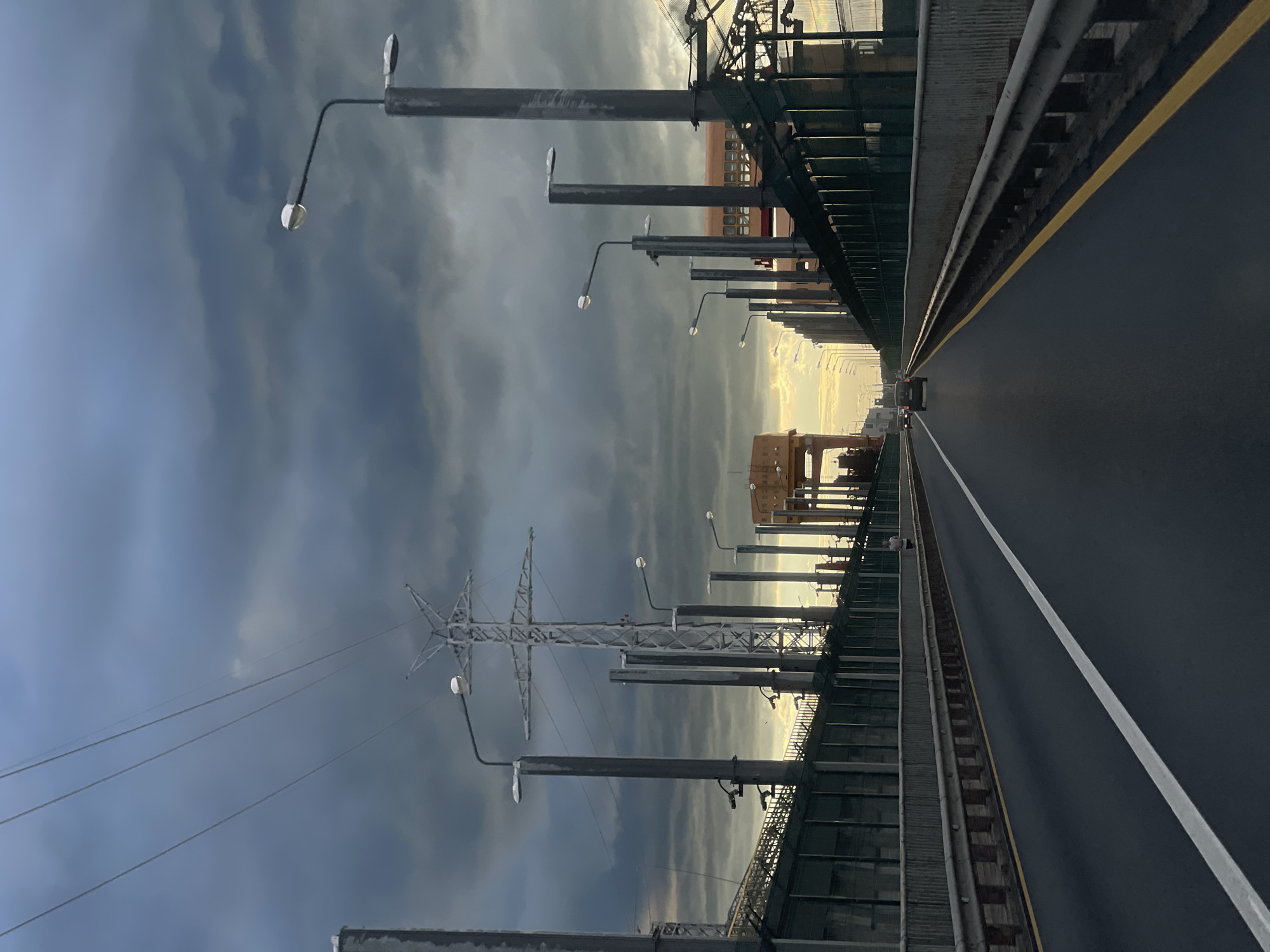 A first-person perspective shot from a vehicle traveling on a road, possibly a bridge, under a dark, cloudy sky. The road is asphalt with a white line dividing the lanes and a yellow line marking the edge. On either side of the road are metal guardrails. Beyond the guardrails, there are tall, dark poles with streetlights. In the background, there are industrial structures, including a large lattice-style power pylon and some yellow buildings. There are a few cars on the road, and one person can be seen walking on the side. The overall atmosphere is somewhat somber due to the overcast weather.